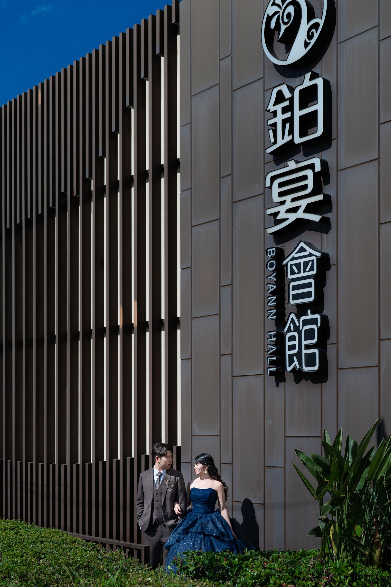 Couple in formal attire posing outside Boyann Hall venue with Chinese signage and modern architectural facade.