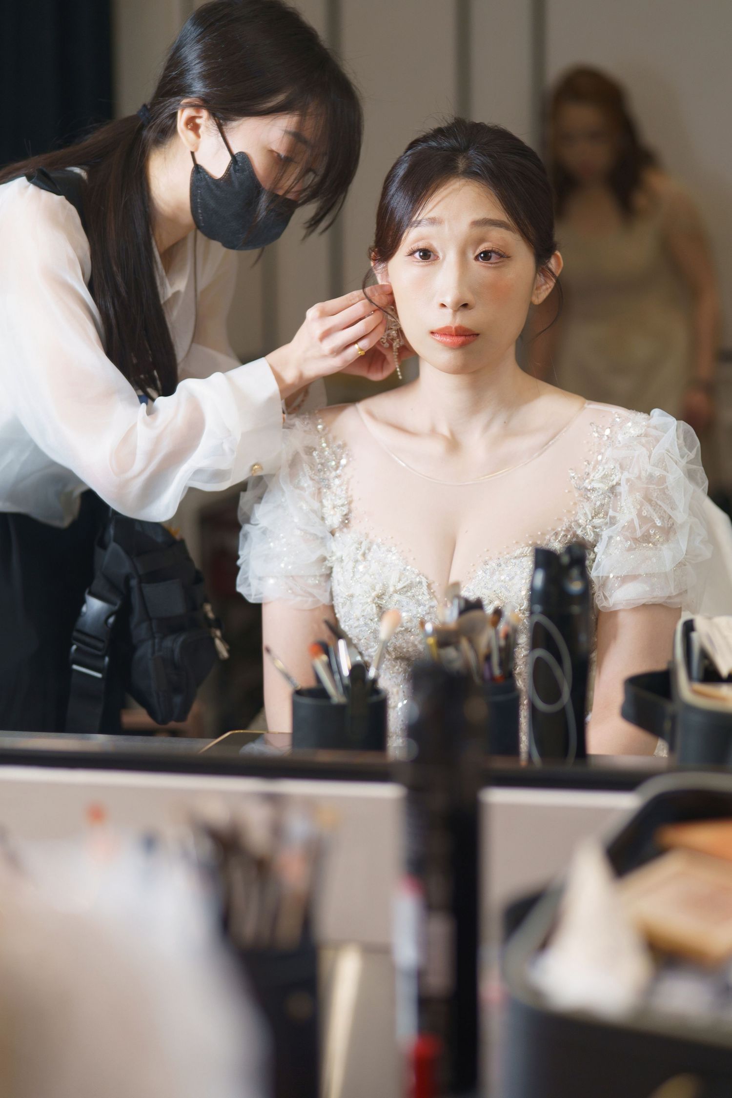 Makeup artist wearing black face mask applies finishing touches to bride's look in white lace dress at beauty station.
