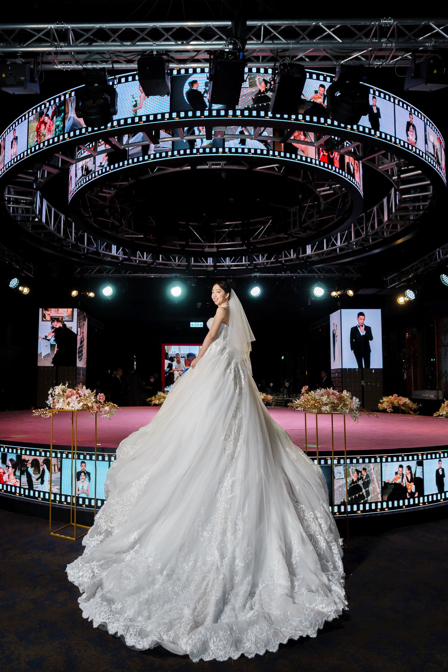 A stunning wedding gown with a long lace train displayed on a circular stage with film strip decorations and video screens.