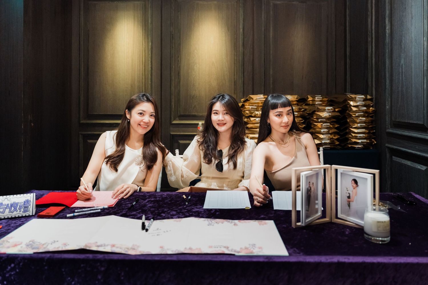 Three people sit at a desk with photo albums and marketing materials displayed on a purple tablecloth.