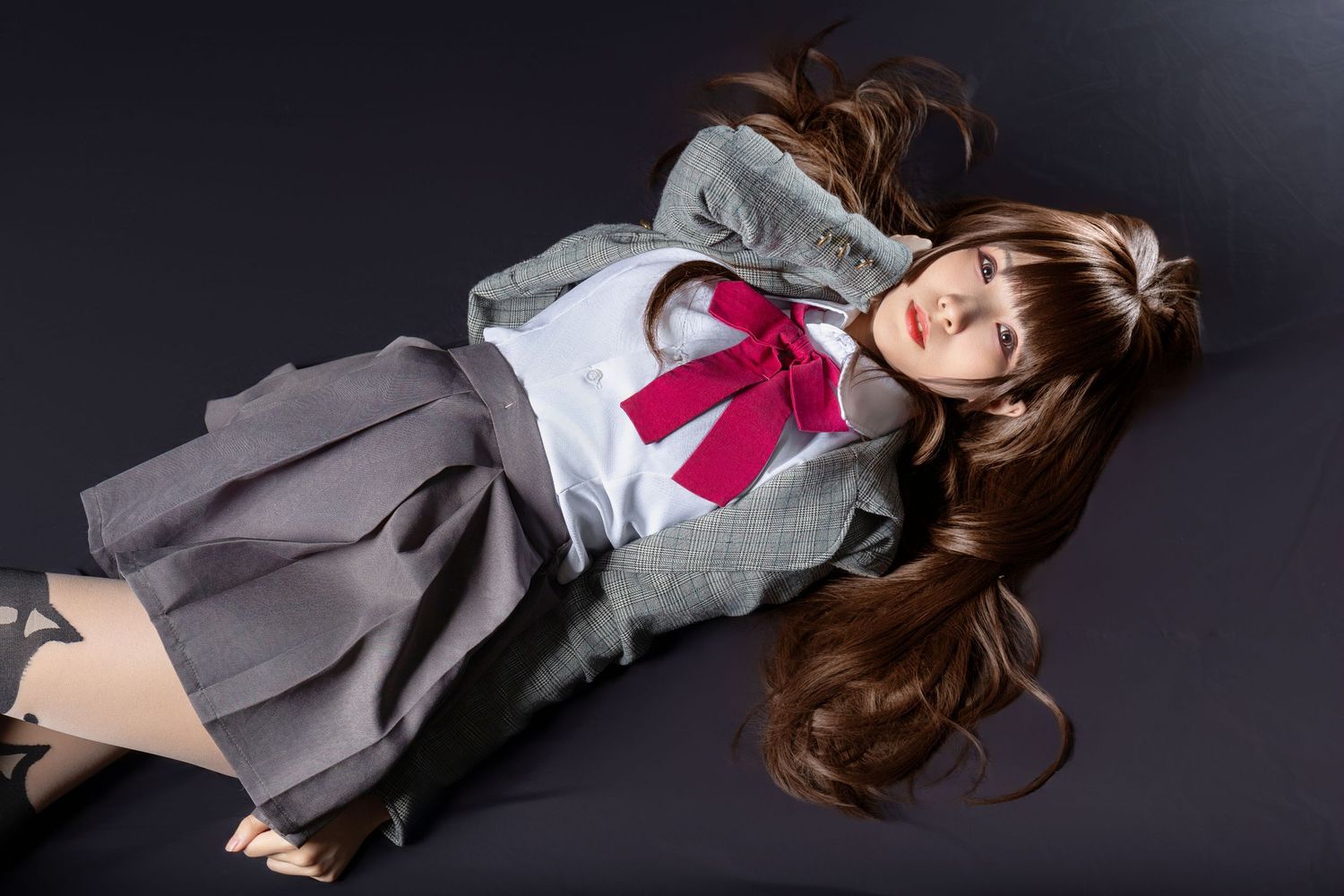 A student in grey uniform with red bow tie lying on dark floor with long hair spread out around them.