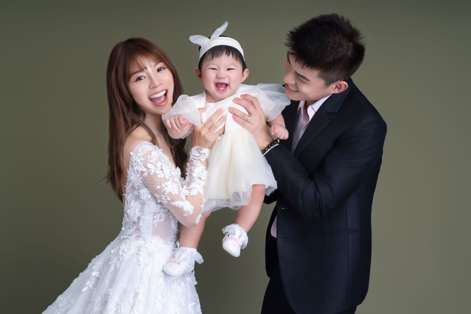 A happy family of three share a joyful moment together against a dark backdrop, dressed in elegant wedding attire.