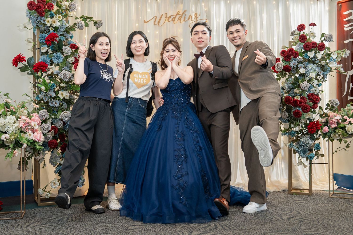 Wedding party in navy blue dresses and suits pose together with red floral arrangements in the background.