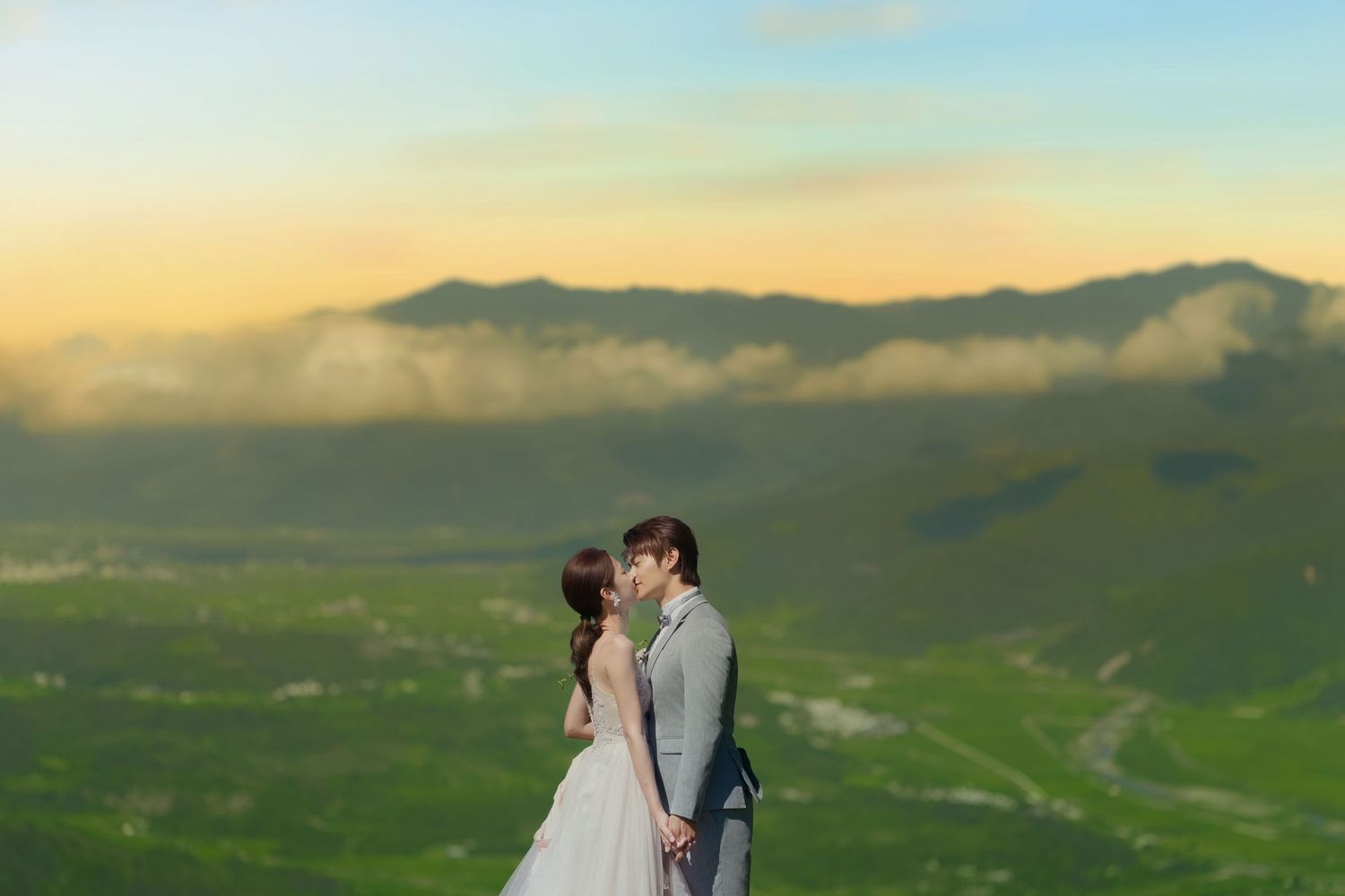 Two people embrace in a romantic moment against a backdrop of green mountainous landscape at sunset.