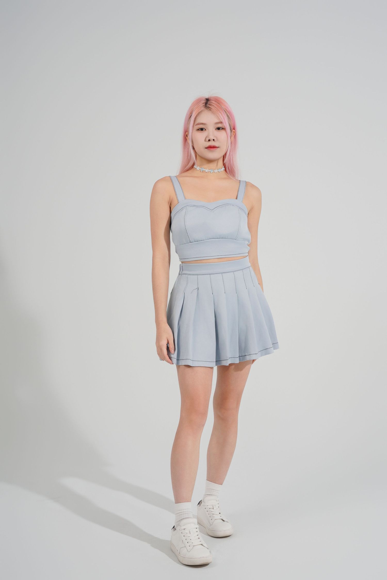 A person in a light blue pleated tennis skirt and crop top poses against a white background wearing white sneakers.