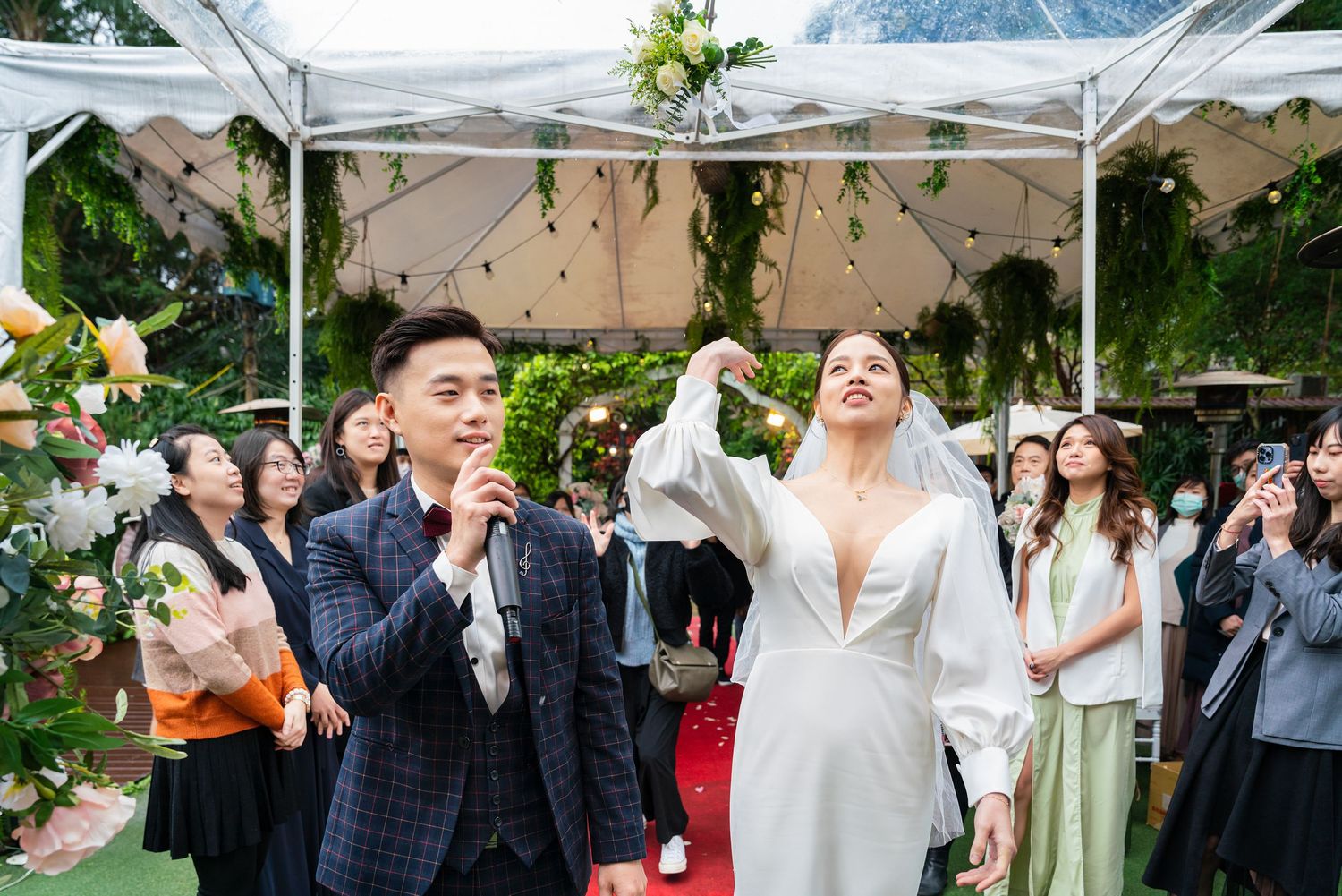 Guests celebrate joyfully at an outdoor garden wedding reception under a white tent decorated with greenery.