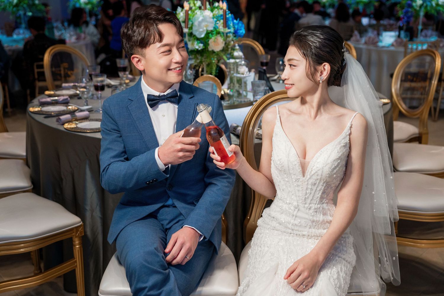 A couple in formal wedding attire sharing a toast at an elegant reception with floral decor.