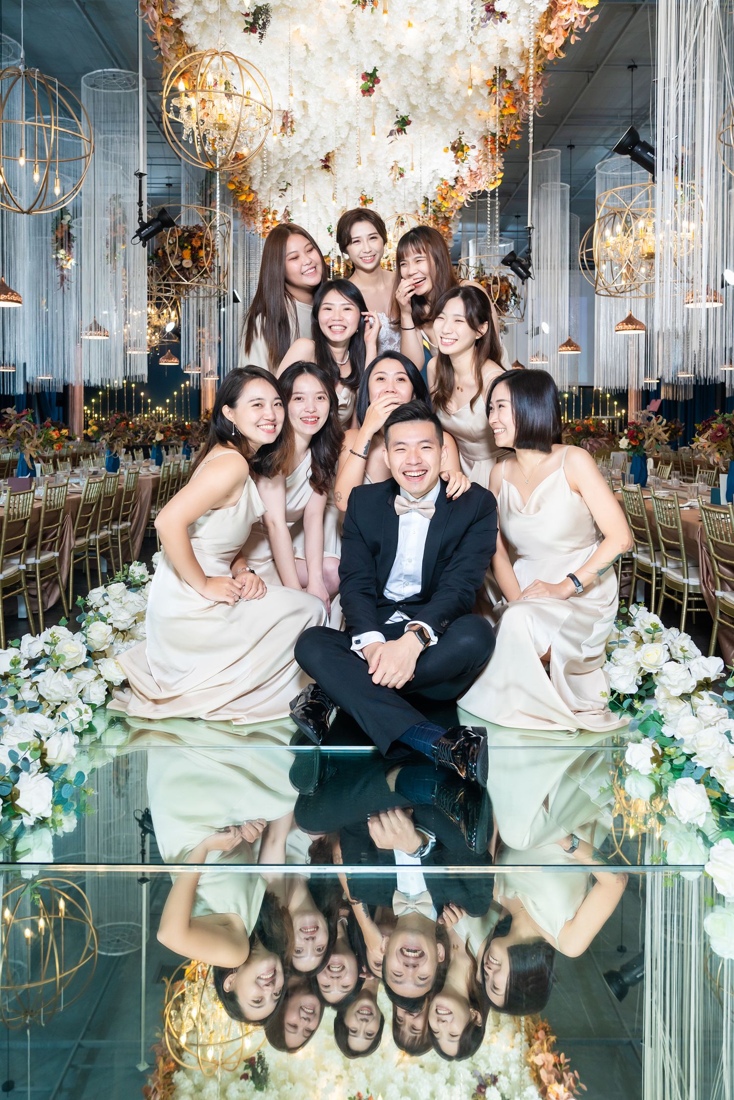 Wedding party group photo in elegant ballroom with mirrored floor and ornate chandelier decorations overhead.
