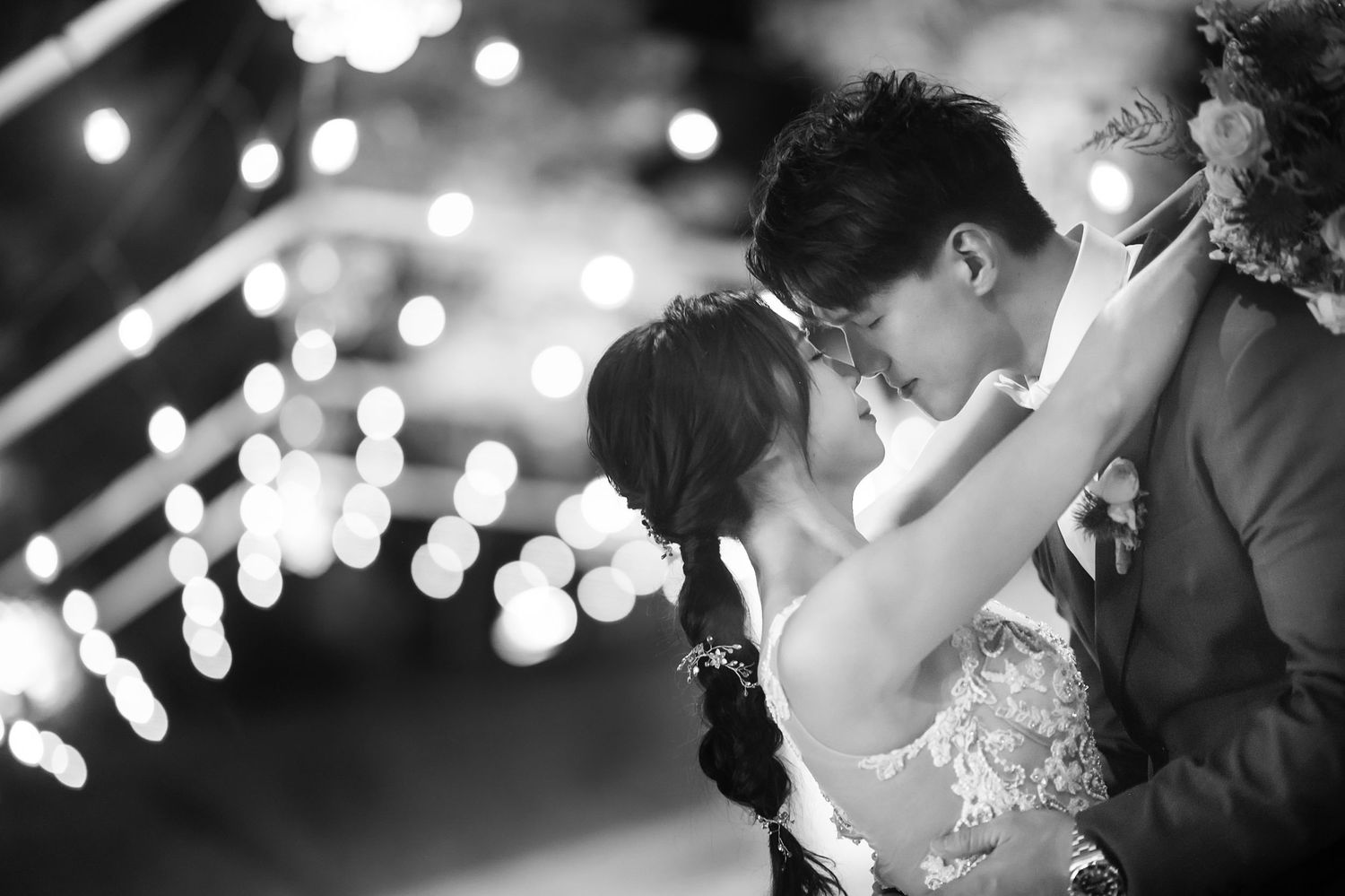A romantic couple shares an intimate moment under twinkling lights at their wedding reception in black and white.