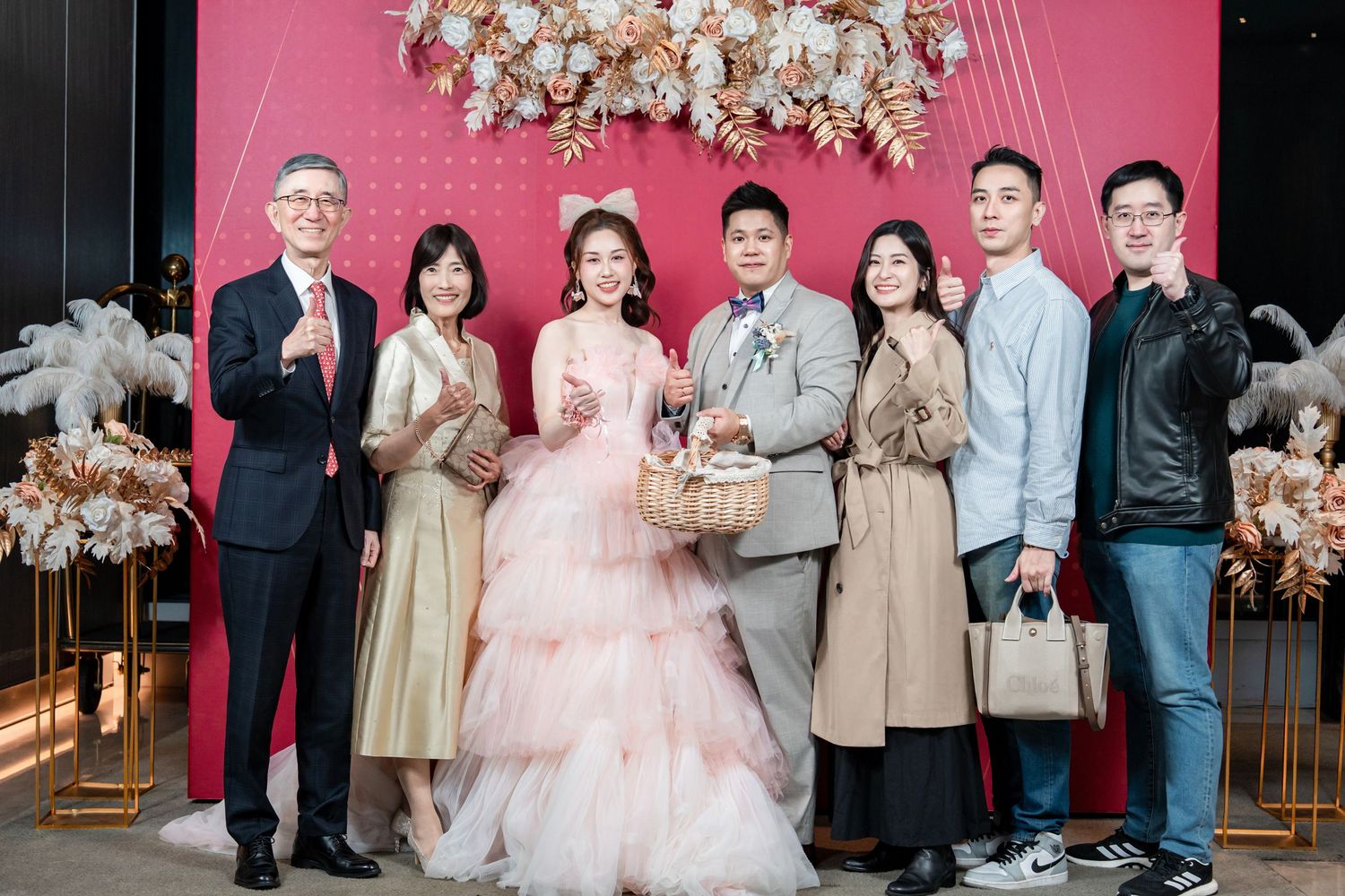 Wedding party stands together against pink backdrop with elegant floral arrangements in formal attire.