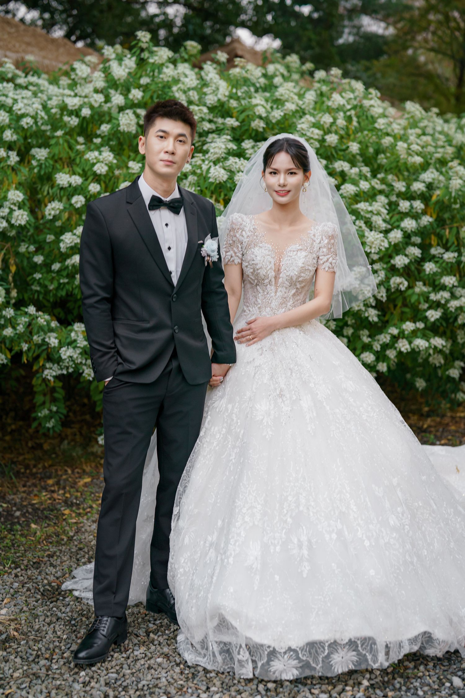 A bride and groom pose together in a garden with blooming white flowers on a spring wedding day.