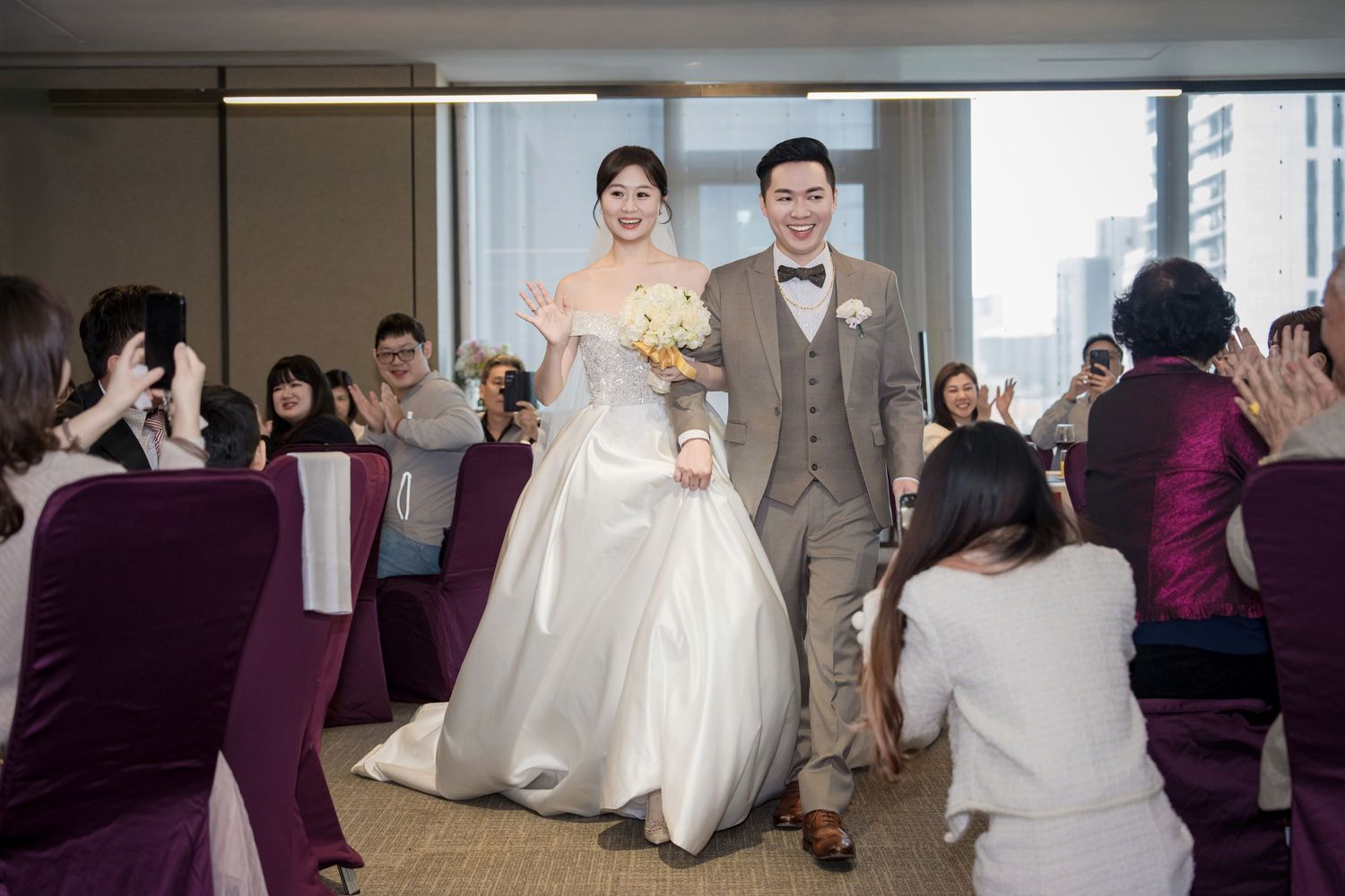 A couple walks down the aisle at their wedding ceremony while guests in purple chairs applaud and take photos.