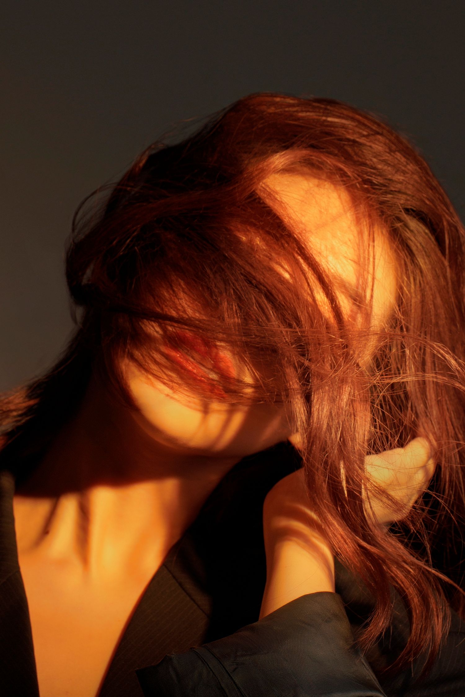 Woman with flowing red hair caught in warm golden light, hand raised, dramatic shadow contrast against dark background.