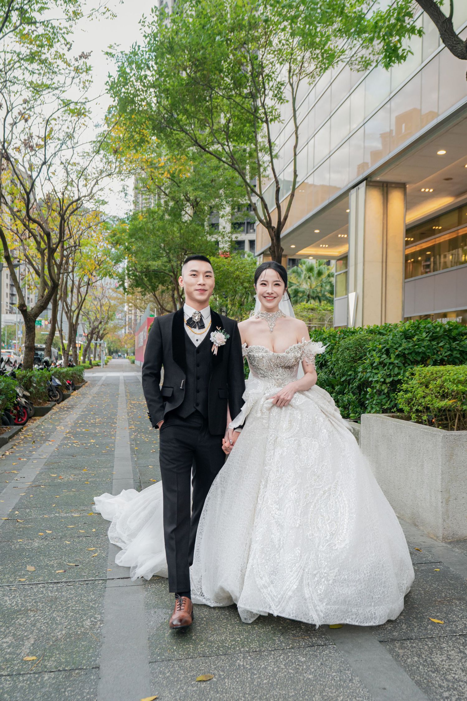 A couple walks down a tree-lined sidewalk outside a modern building, with the bride wearing a sparkly ballgown.
