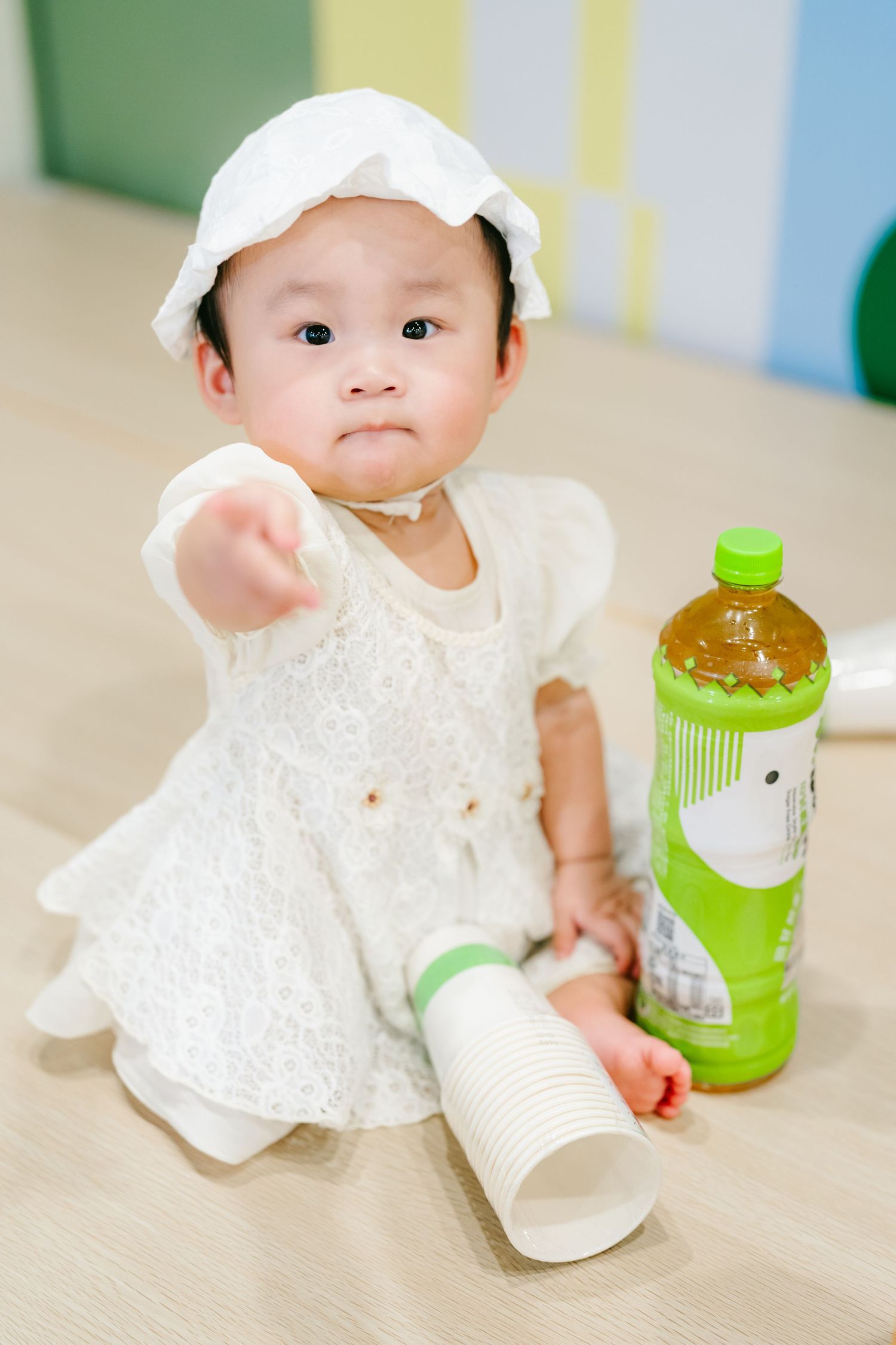 Small child in white dress and cap sits on floor with green tea bottle and paper cup.