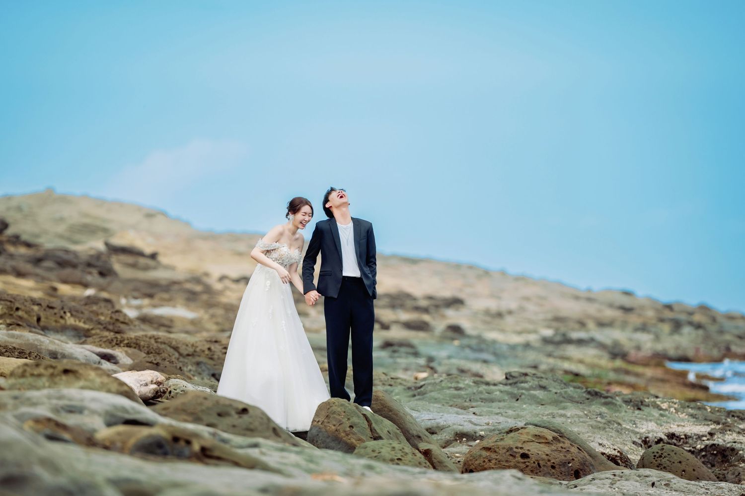 A couple in formal wedding attire stands together on rocky coastal terrain against a bright blue sky.