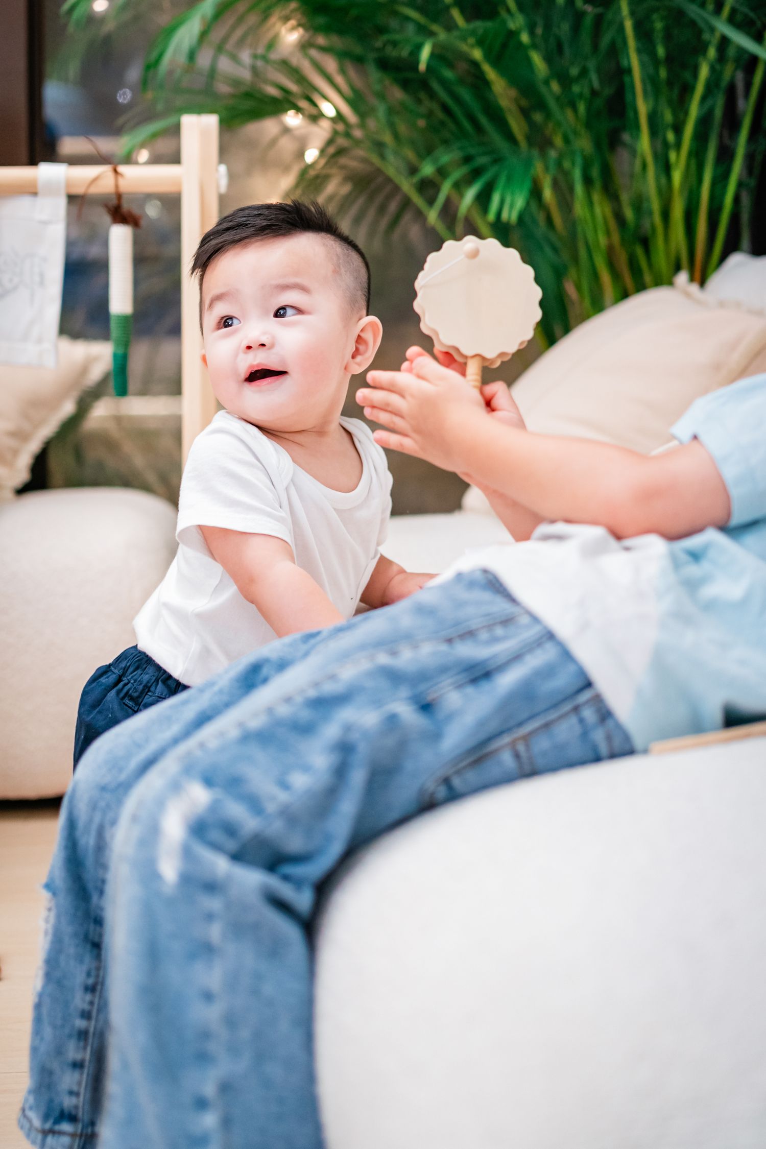 A small child in a white shirt sits on a couch while someone holds up a toy in the background.