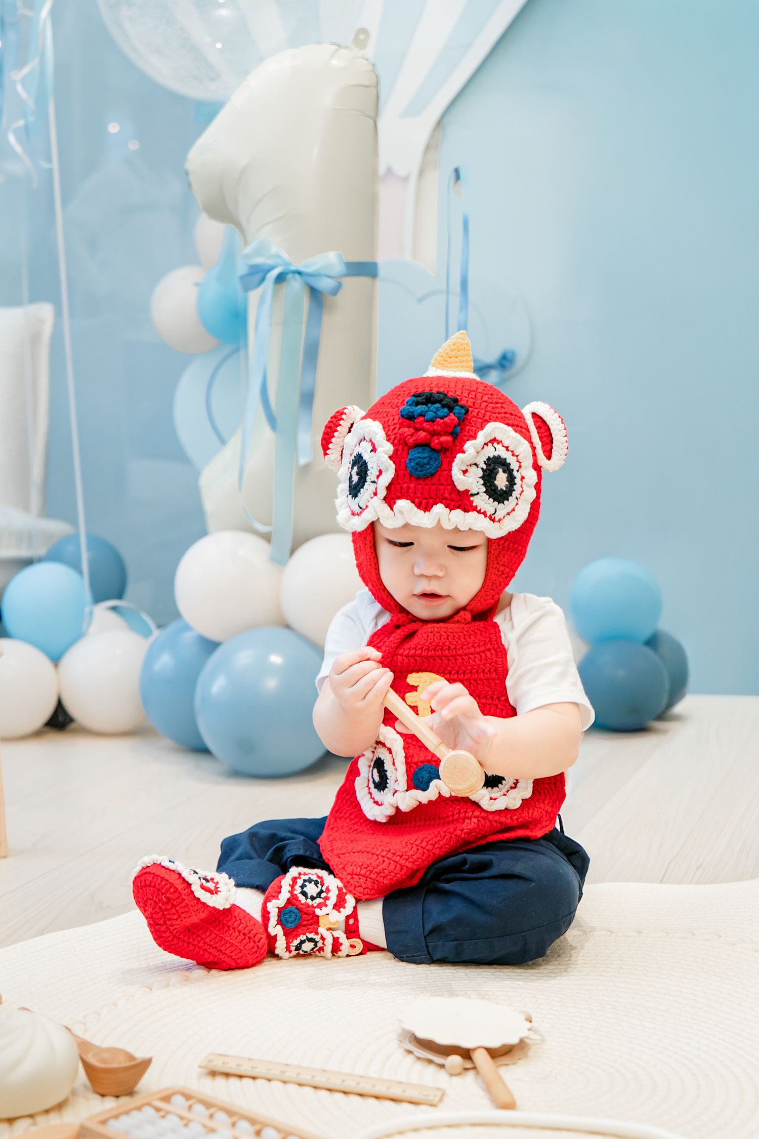A baby in a red knit crab costume sits on a white floor surrounded by blue balloons and party decorations.