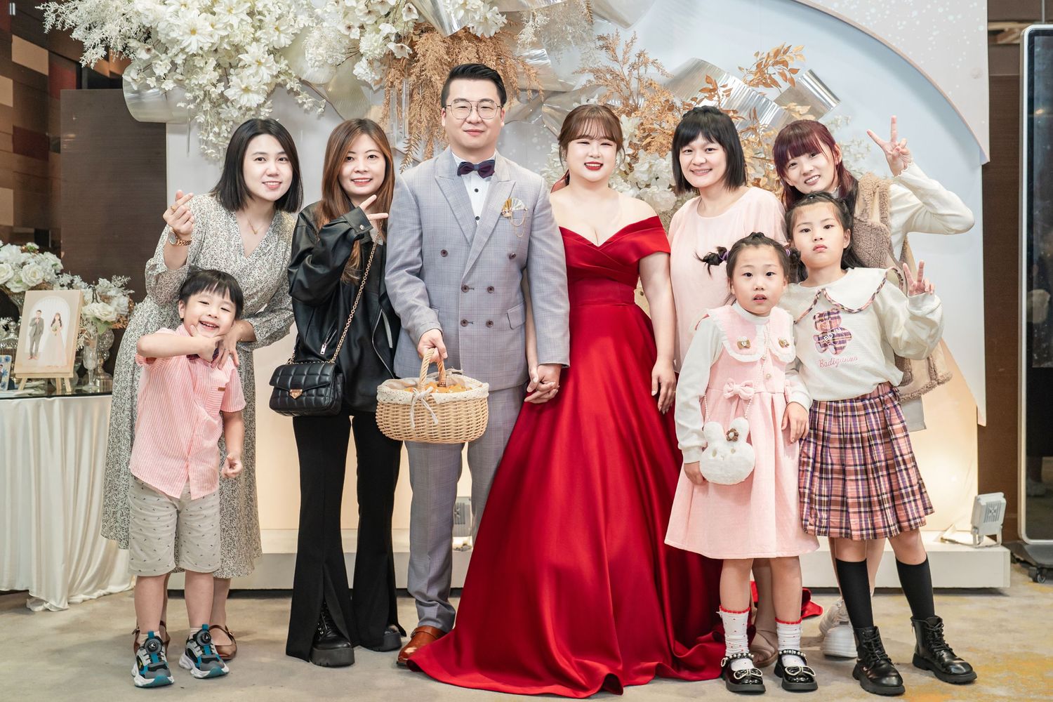 Children in formal attire stand together at an elegant event with white floral decorations in the background.