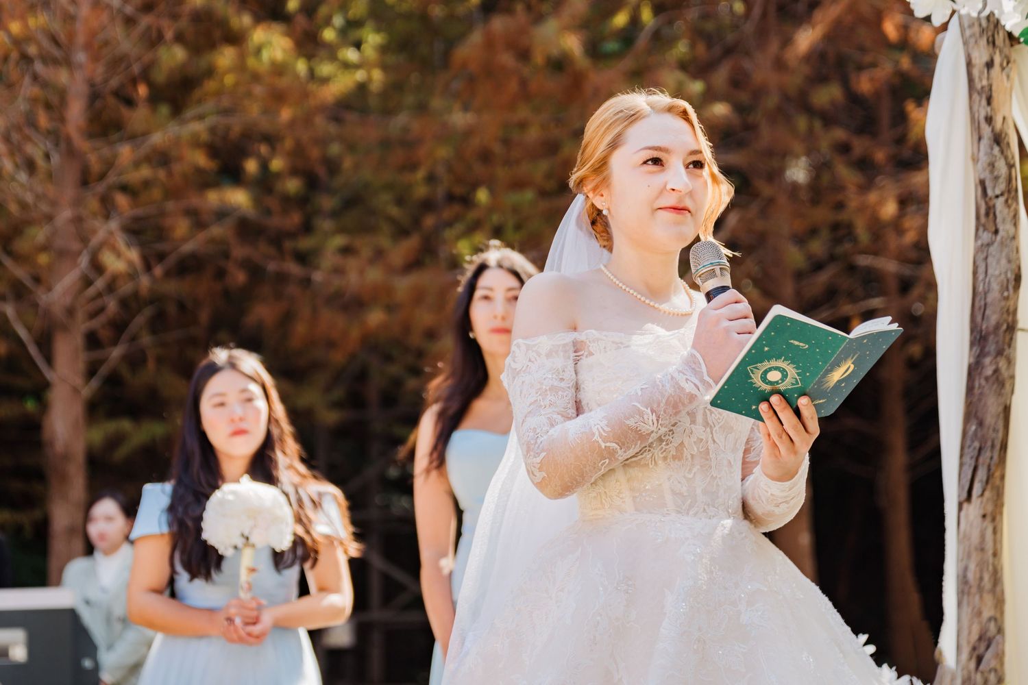 A bride in lace gown holds a book during an outdoor autumn ceremony.