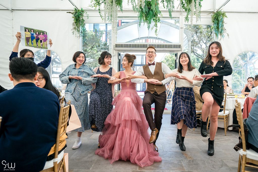 Wedding party dances in a line at reception venue decorated with hanging greenery.