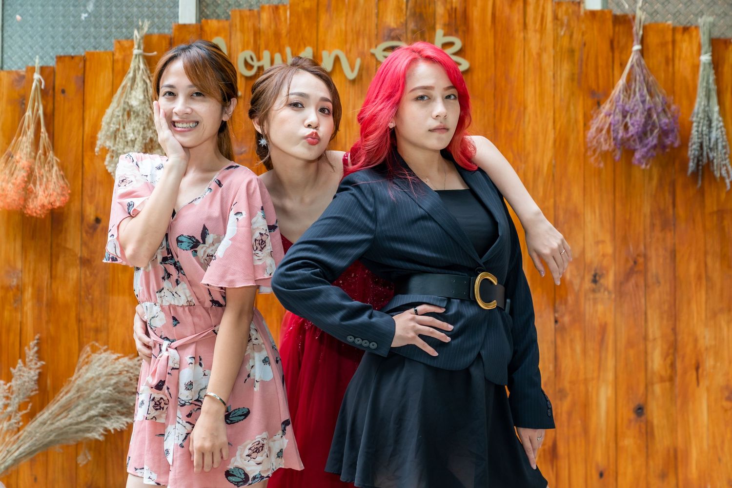 Three models in vintage-inspired dresses pose together against an orange wooden wall with dried flowers.