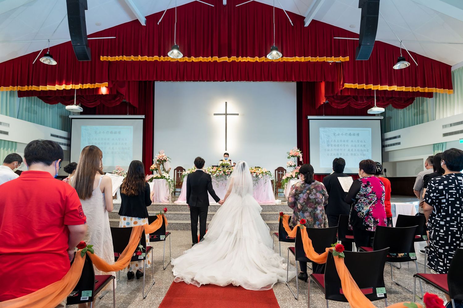 Wedding ceremony in a church with red velvet curtains and illuminated cross at the altar.