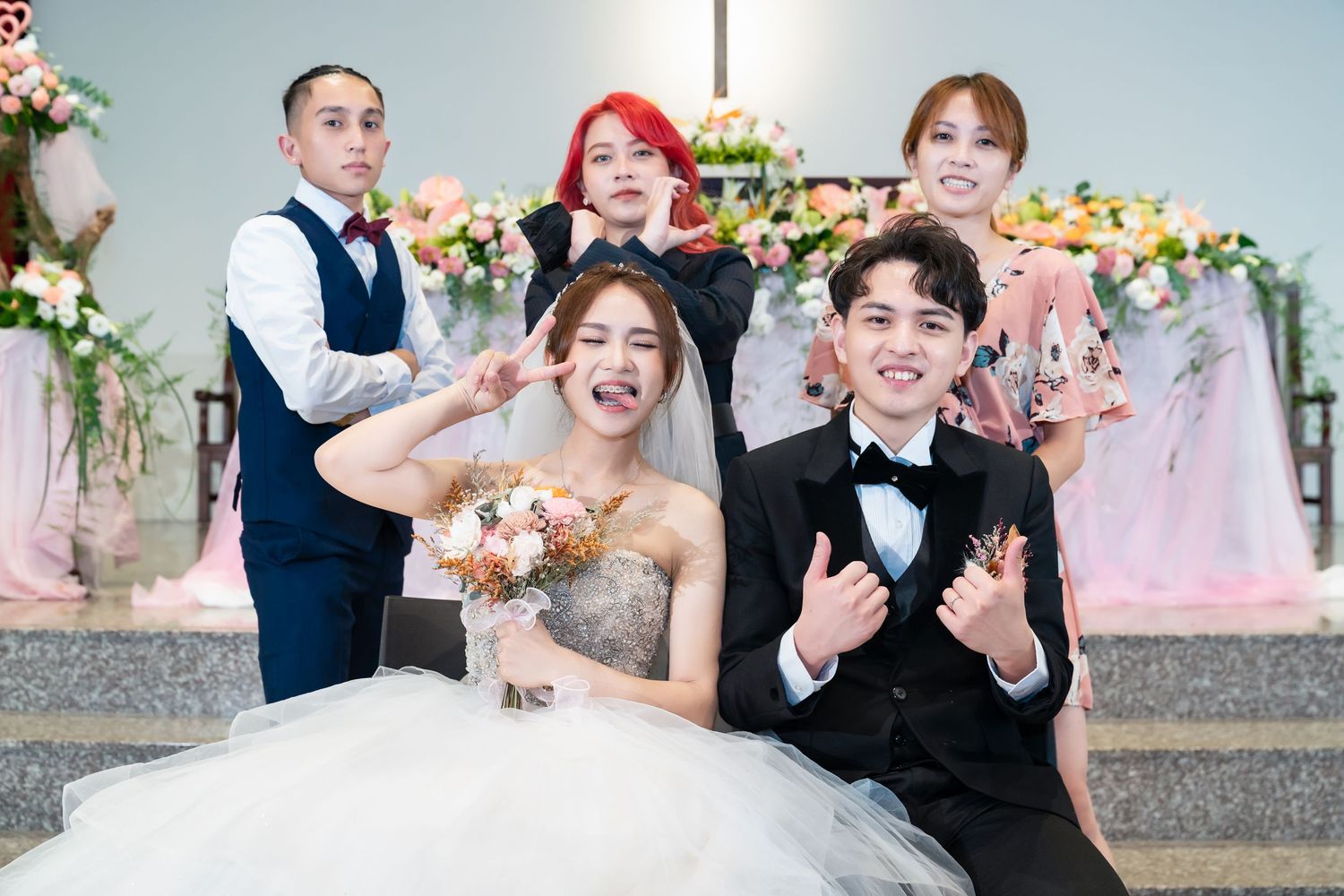 Wedding party poses playfully on church steps with floral arch and decorative backdrop.