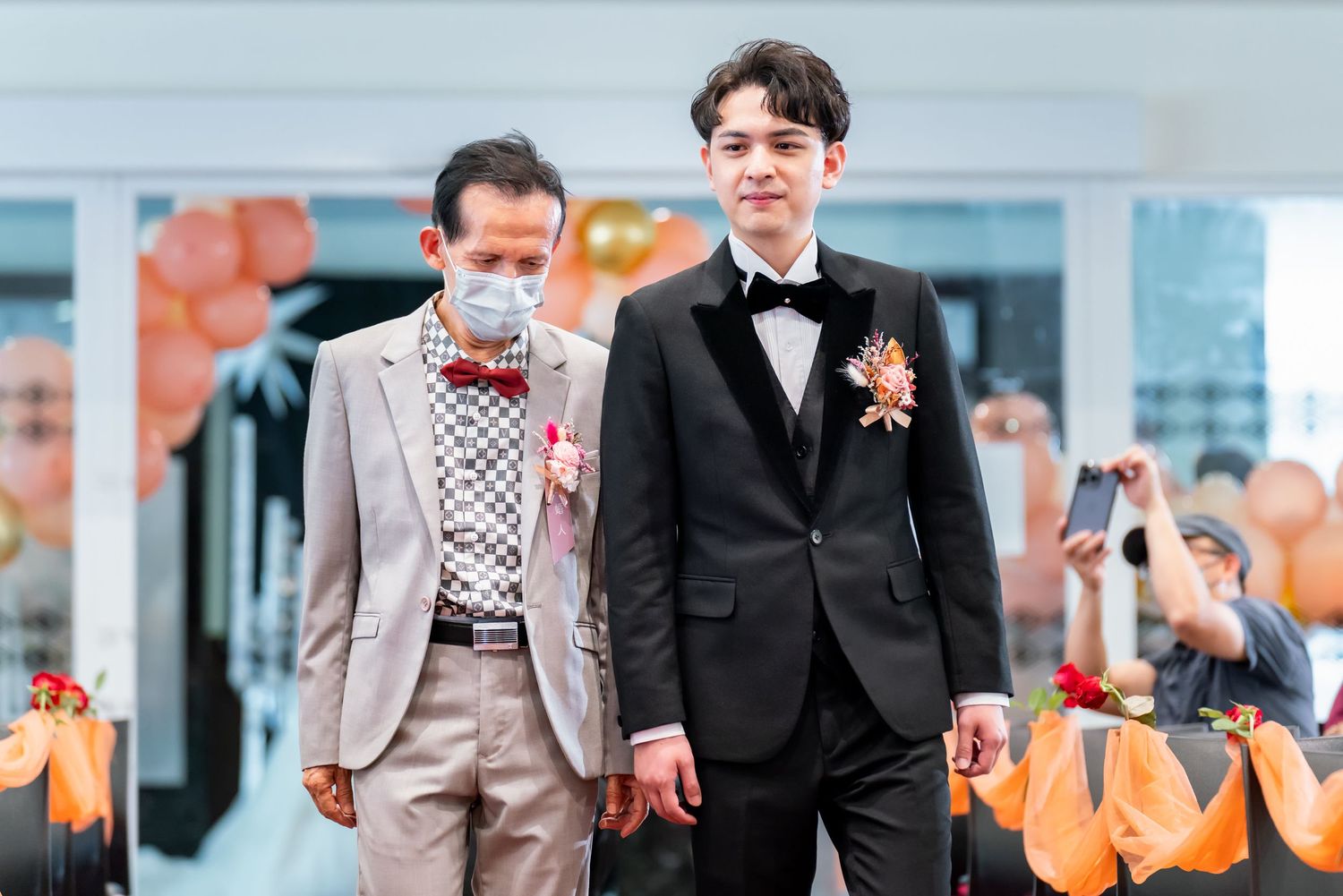 Two formally dressed guests in suits and face masks at wedding celebration.