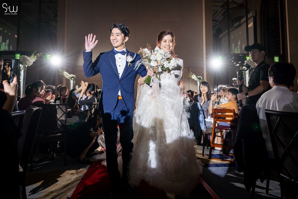 Wedding couple waves to guests during reception in elegant venue with dramatic lighting.