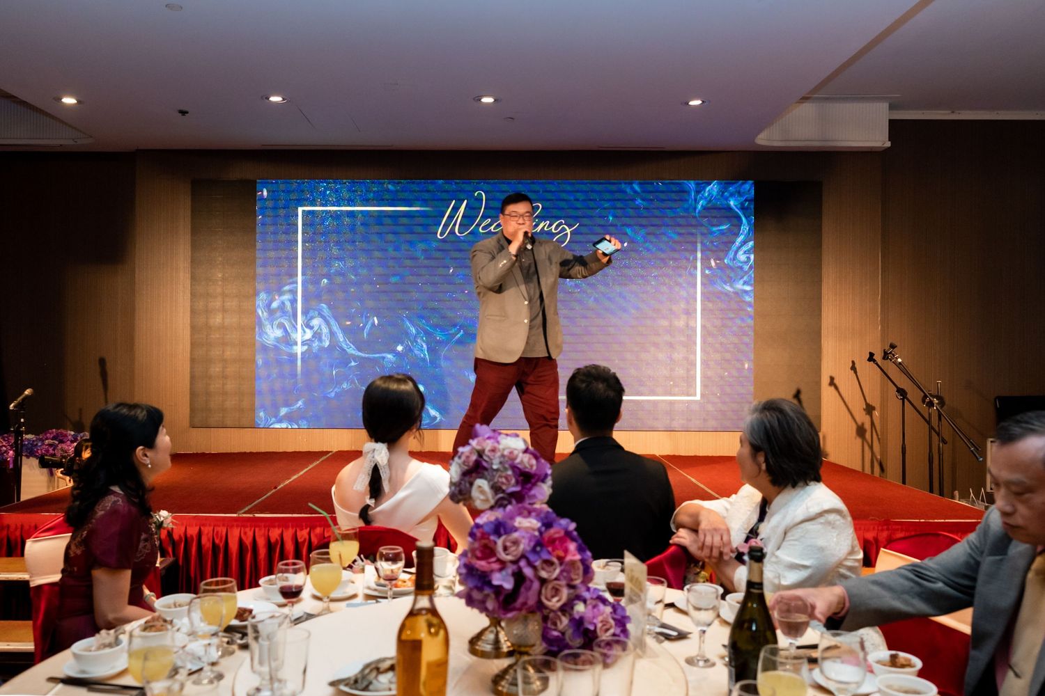 A performer entertains wedding guests during dinner with purple floral centerpieces visible.