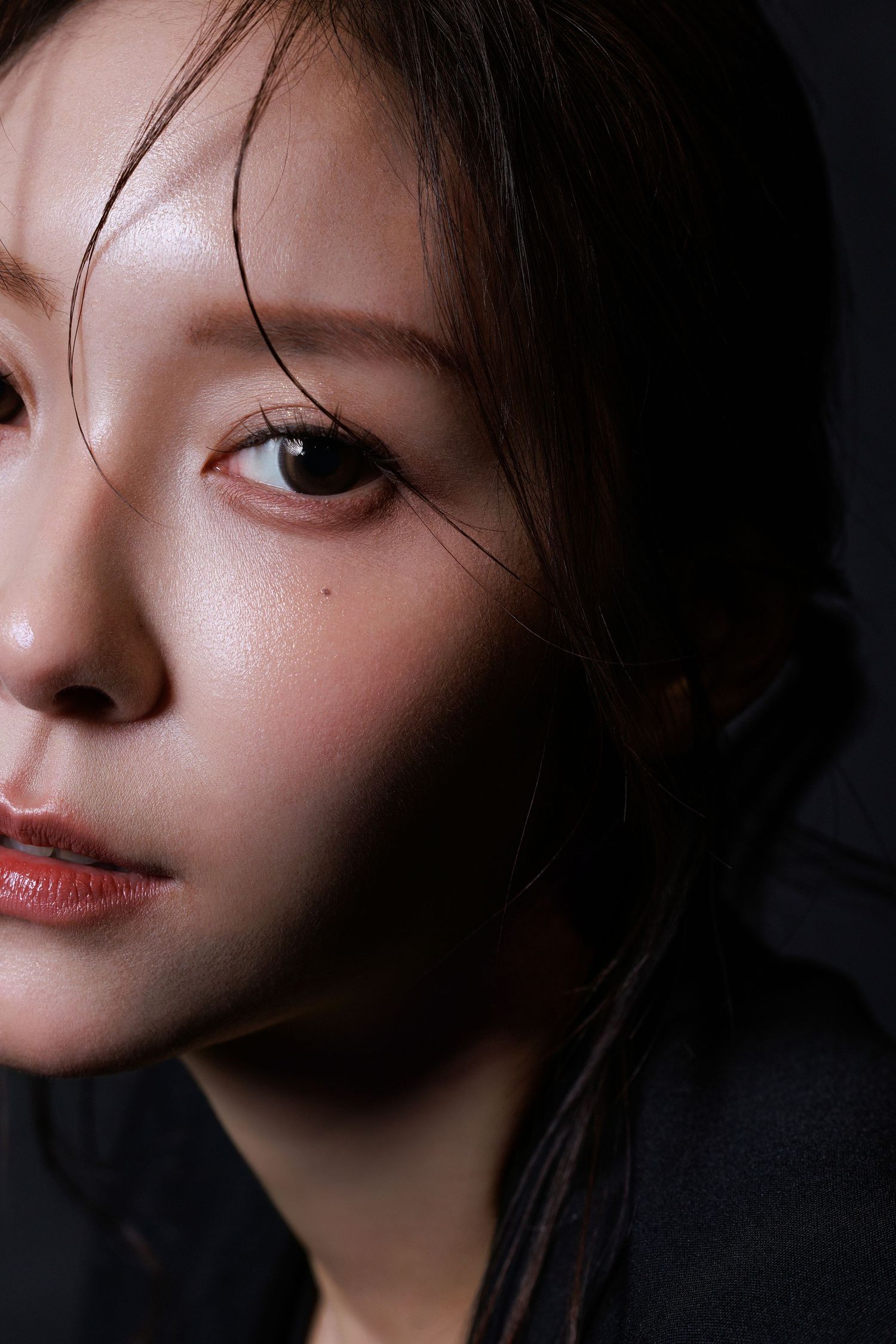 Close-up portrait of a young Asian woman with dark hair, subtle makeup, and a contemplative expression against a dark background.