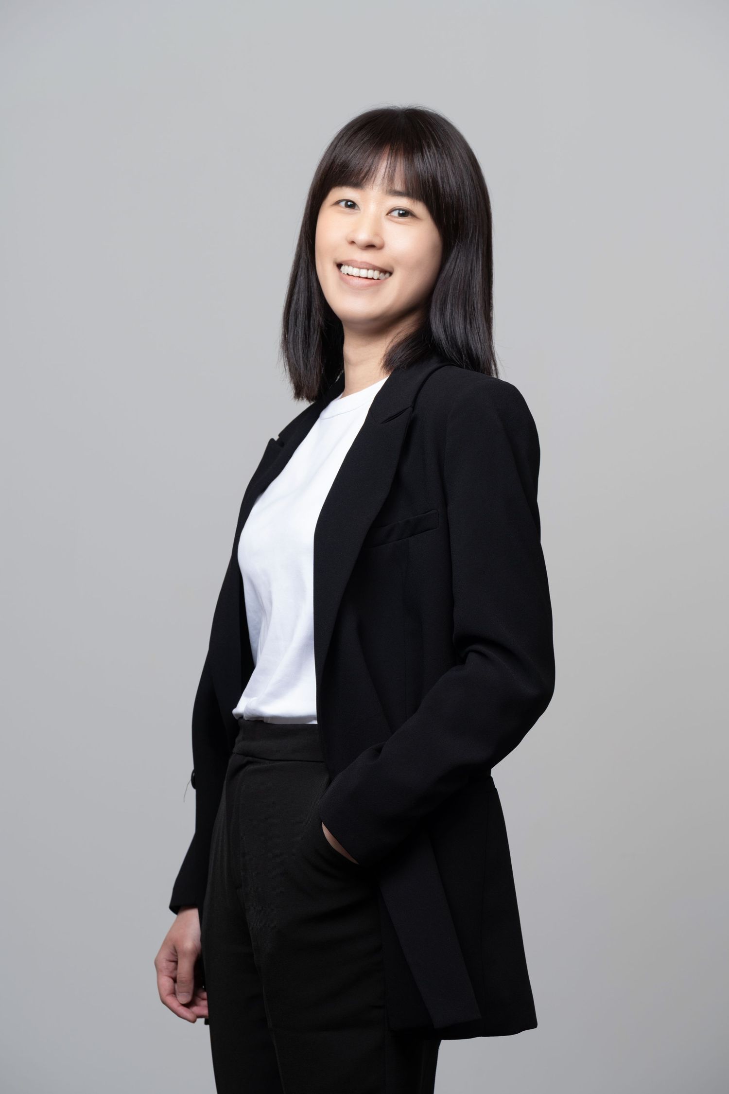 Professional business portrait showing someone in a polished black blazer and white shirt smiling against a gray background.