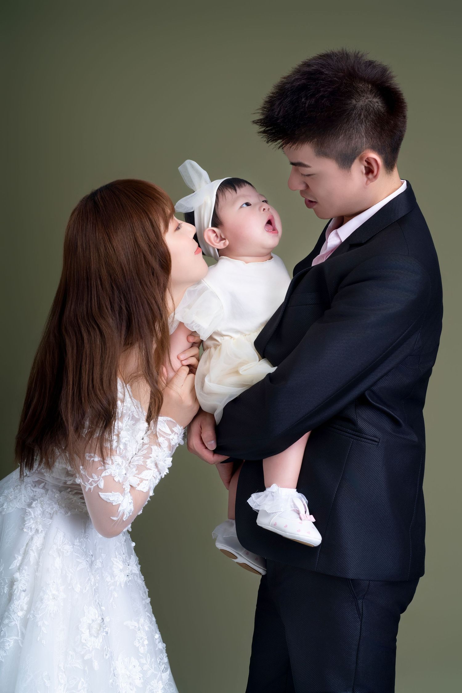 A family portrait showing parents in formal wedding attire holding their baby in white against a green backdrop.