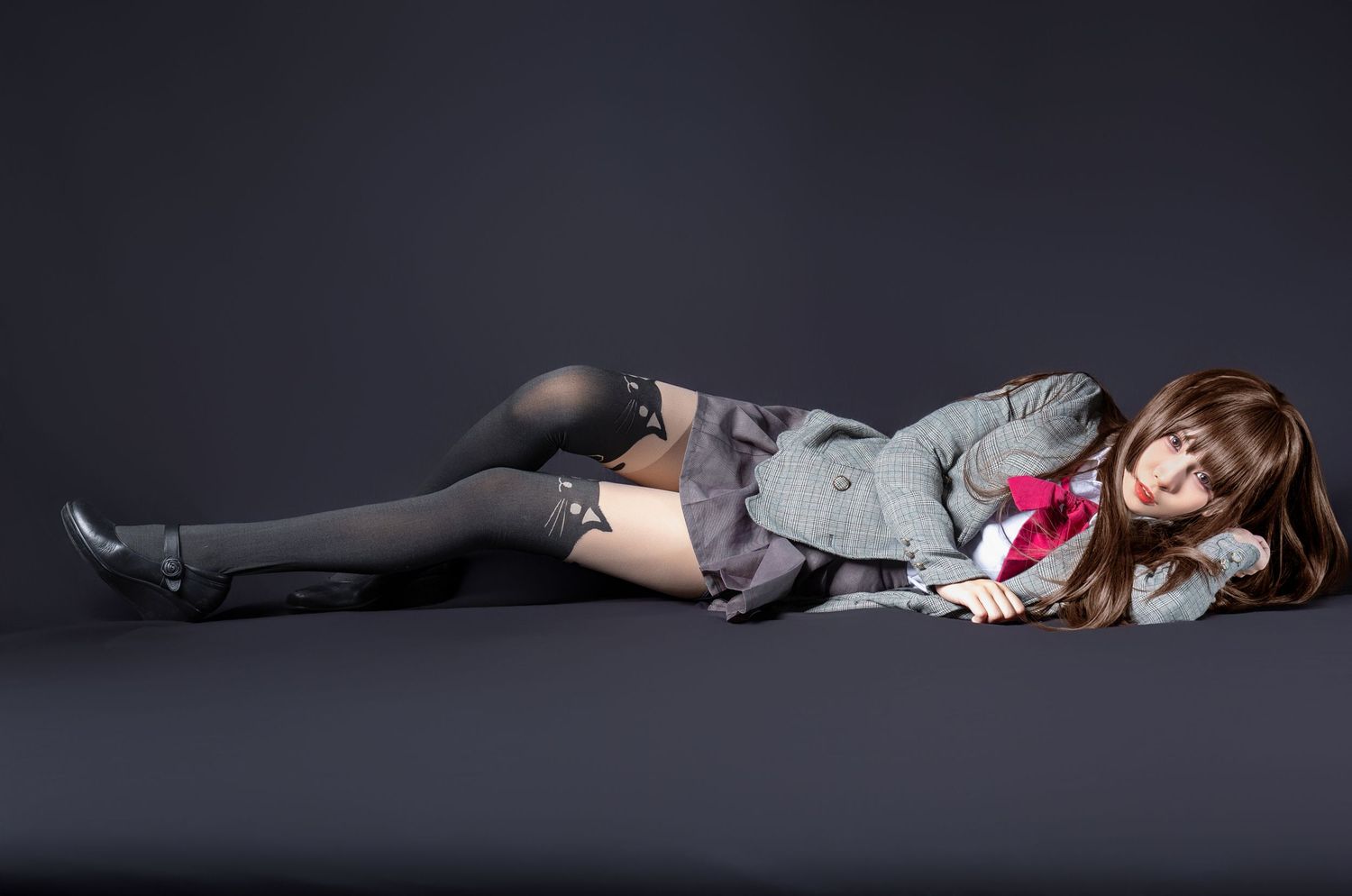 A figure lying on grey floor wearing knee-high stockings, shorts and a cardigan in moody dark studio lighting.