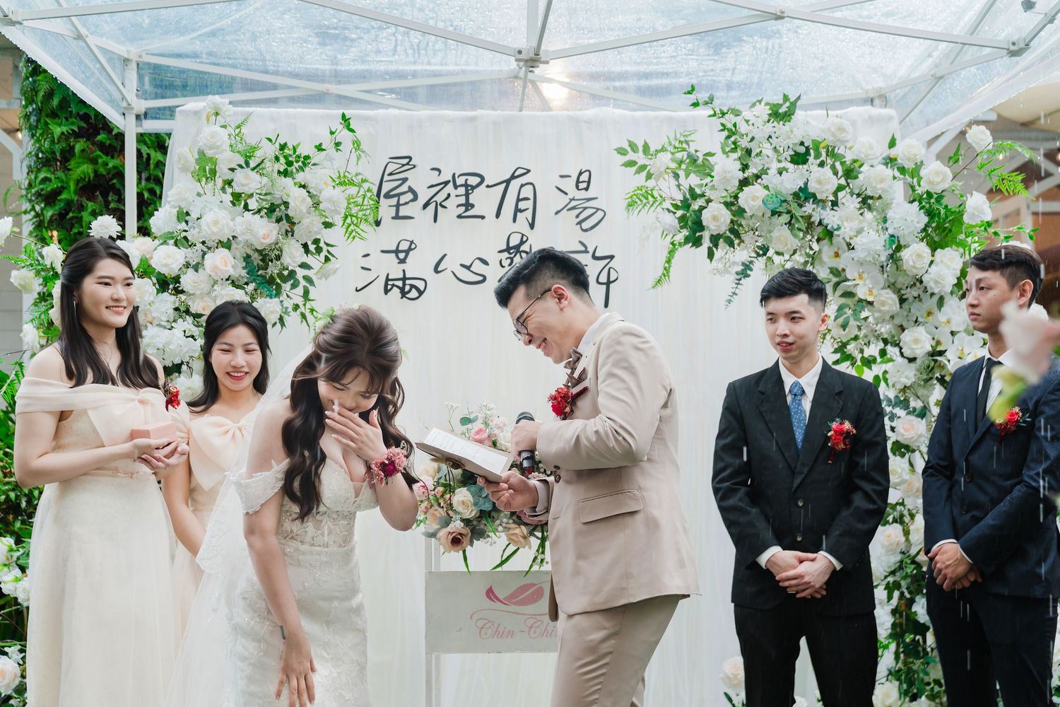 A wedding ceremony taking place under a white floral arch with bridesmaids in light dresses and groomsmen in dark suits.