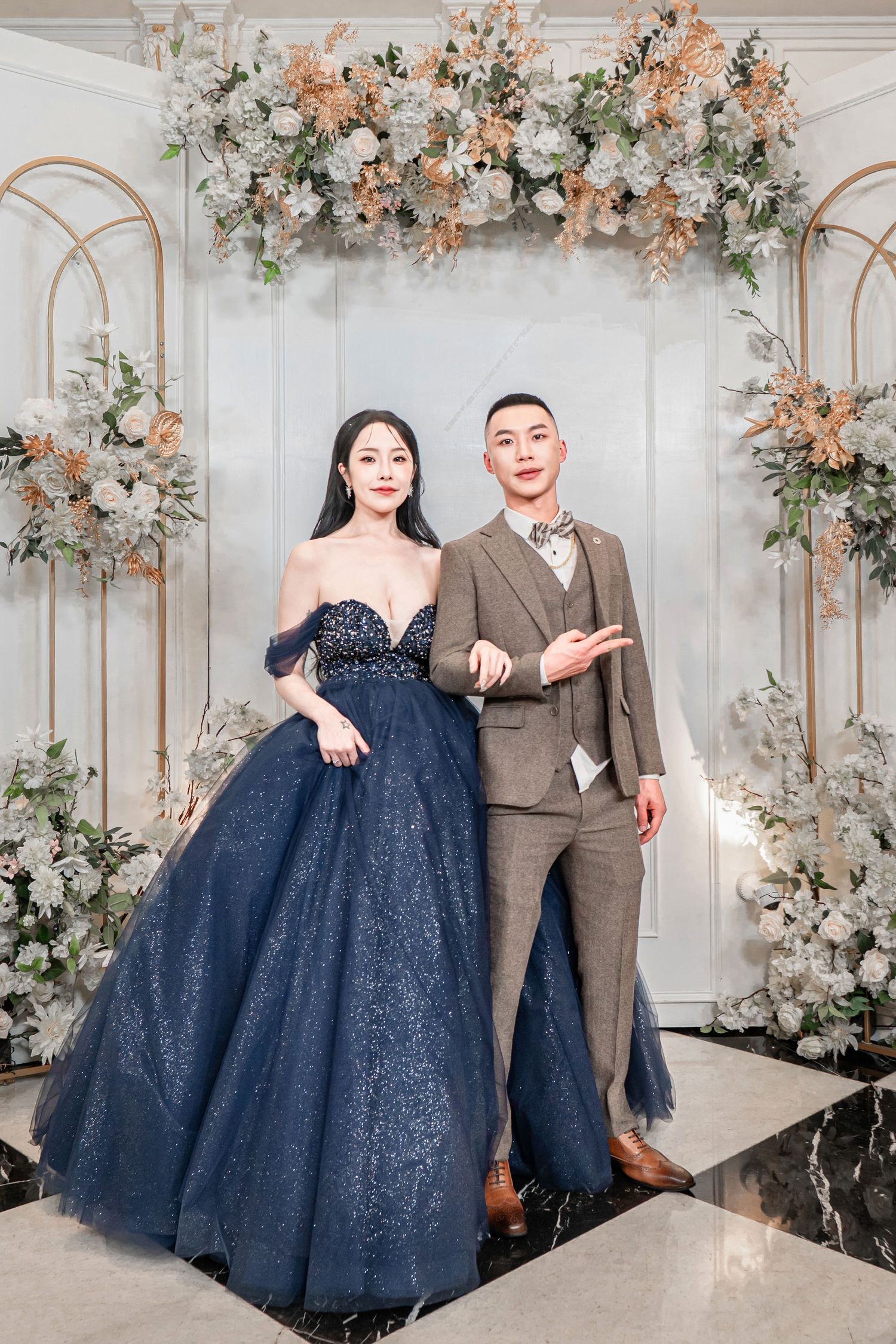 Elegant navy blue ball gown and brown suit in a romantic floral-decorated white interior setting for wedding photos.