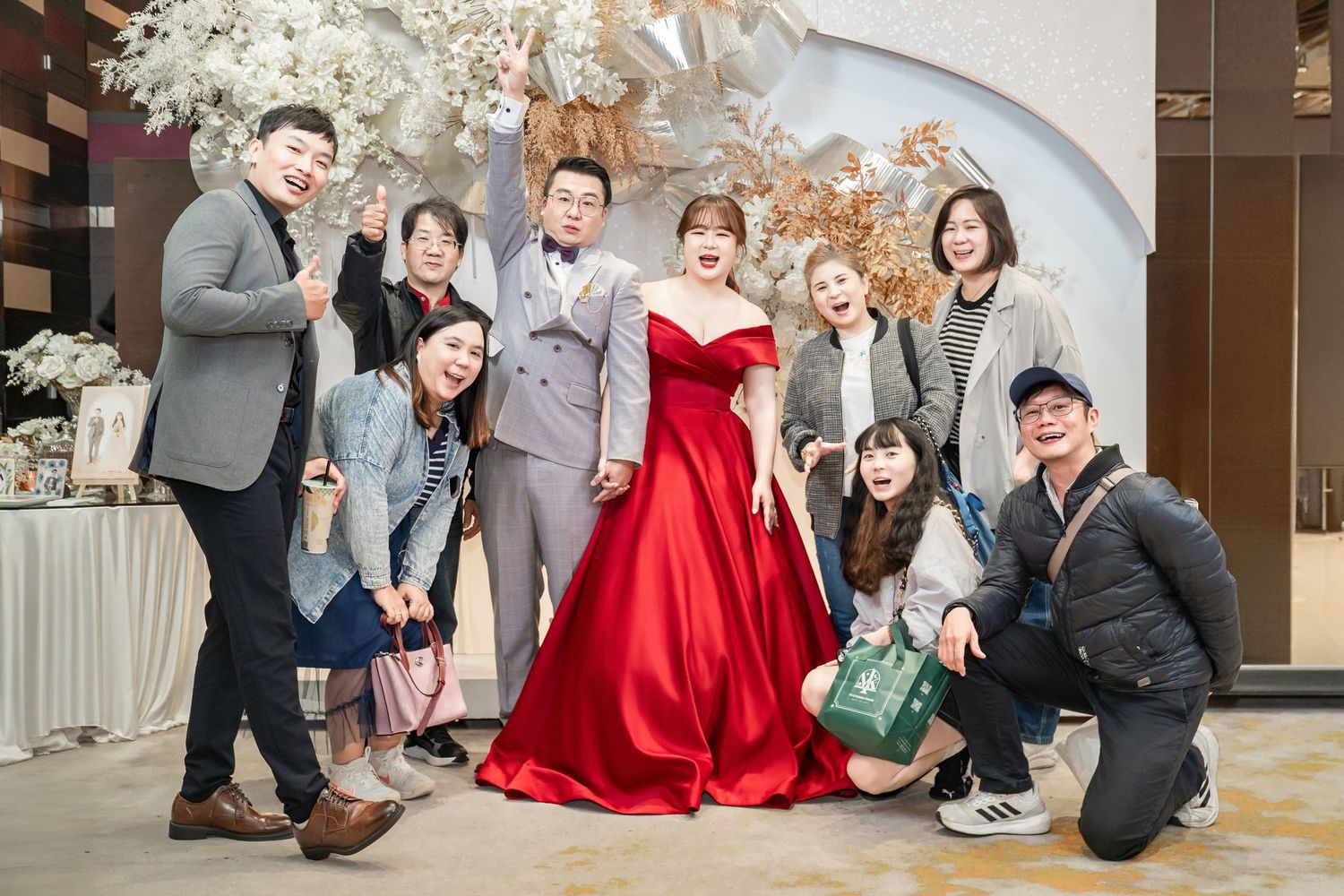 A group of wedding guests pose together during a celebration, with some wearing formal attire and red dresses.
