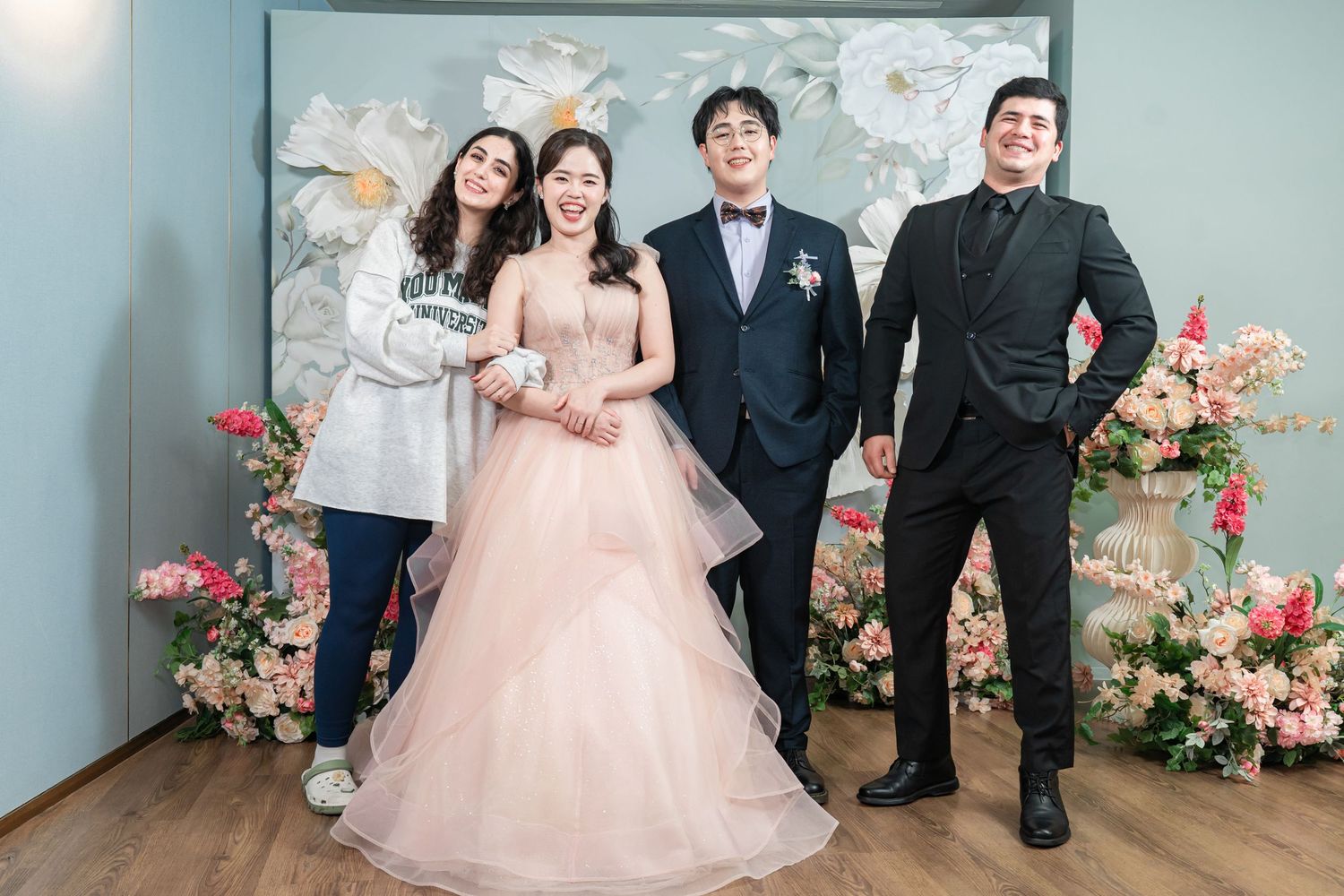 Wedding party poses together in formal attire with pink floral decorations and light blue walls in the background.