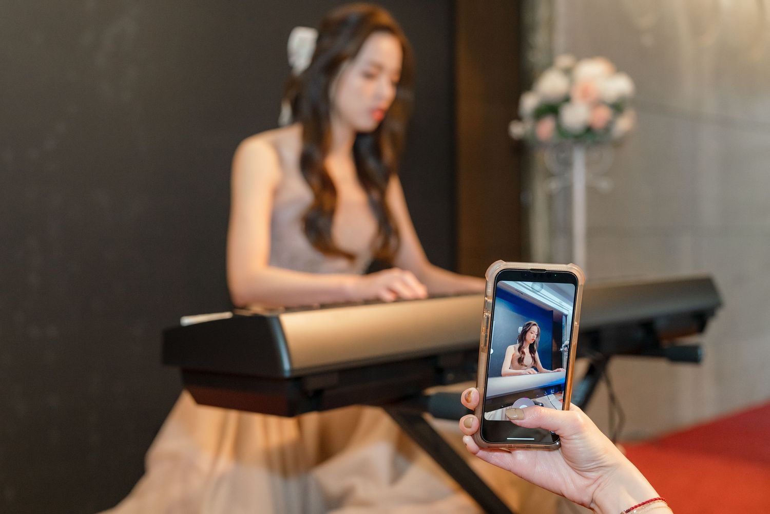 A pianist in a flowing dress sits at a keyboard while someone holds up a smartphone displaying a photo.