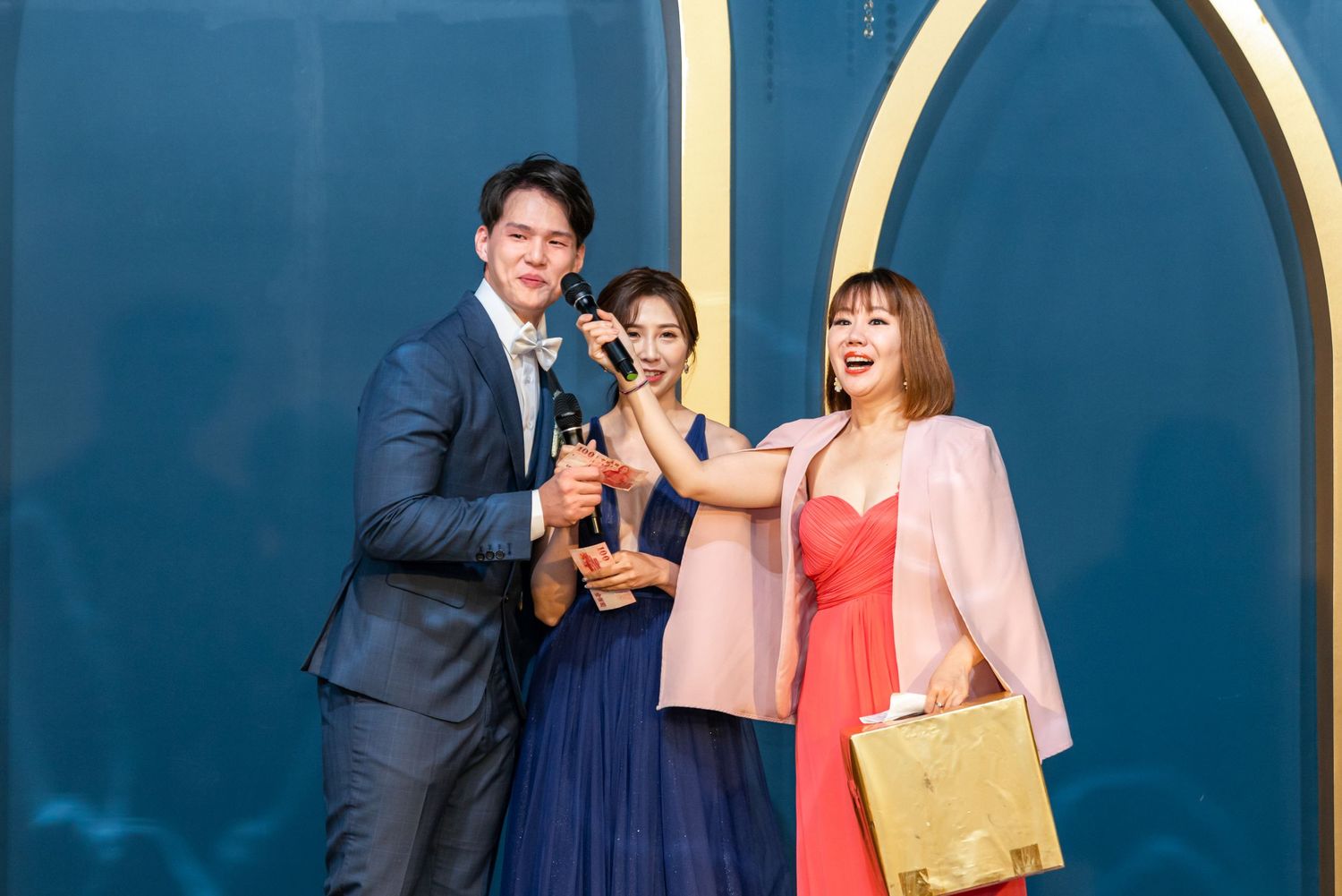 Three people pose together in formal attire against a blue and gold backdrop at an event.