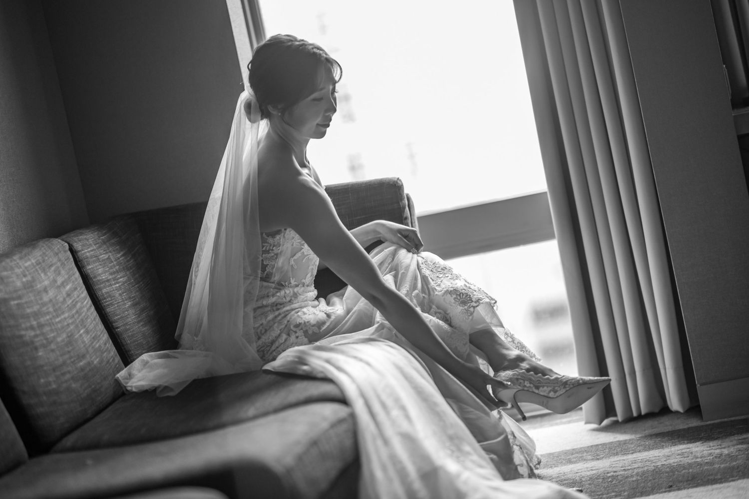 A bride in an elegant white dress sits by a window while putting on her shoes in black and white photography.