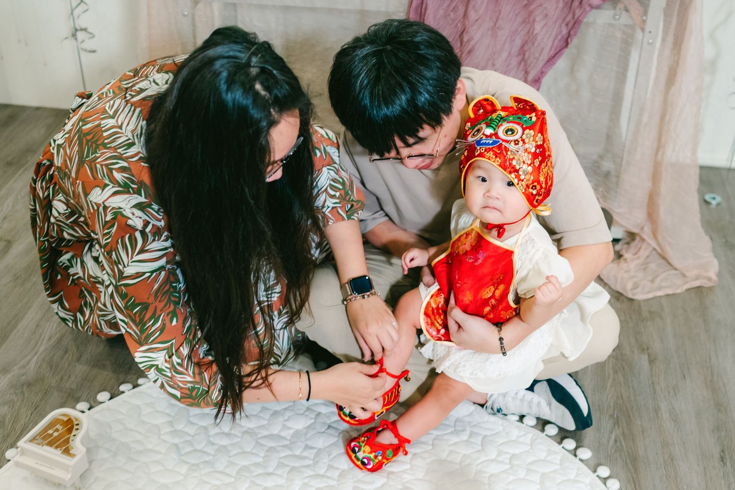 Parents help a baby in a festive red traditional Chinese outfit put on matching red shoes while sitting on the floor.