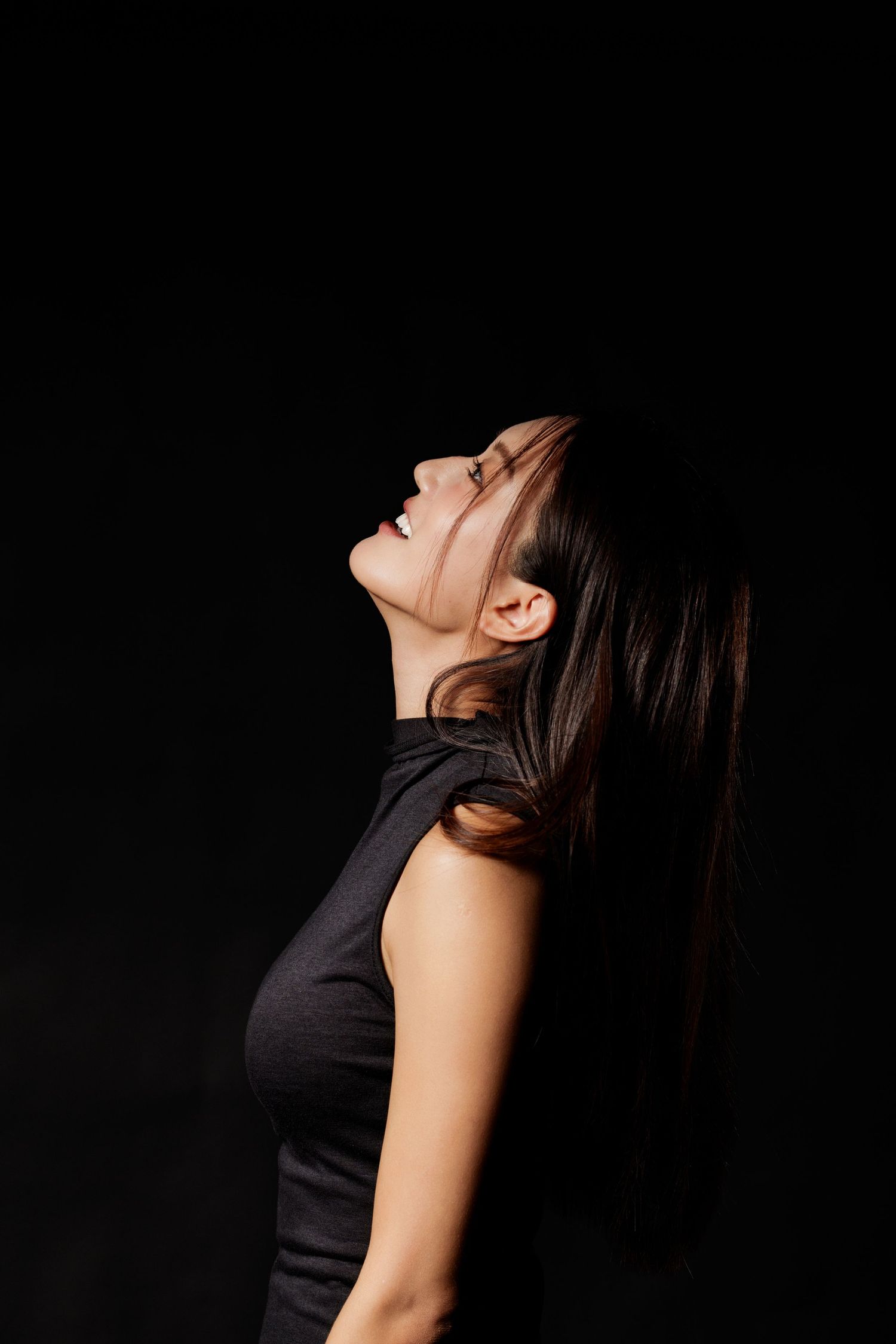 Young woman in black sleeveless top laughing with head tilted back against dark background.