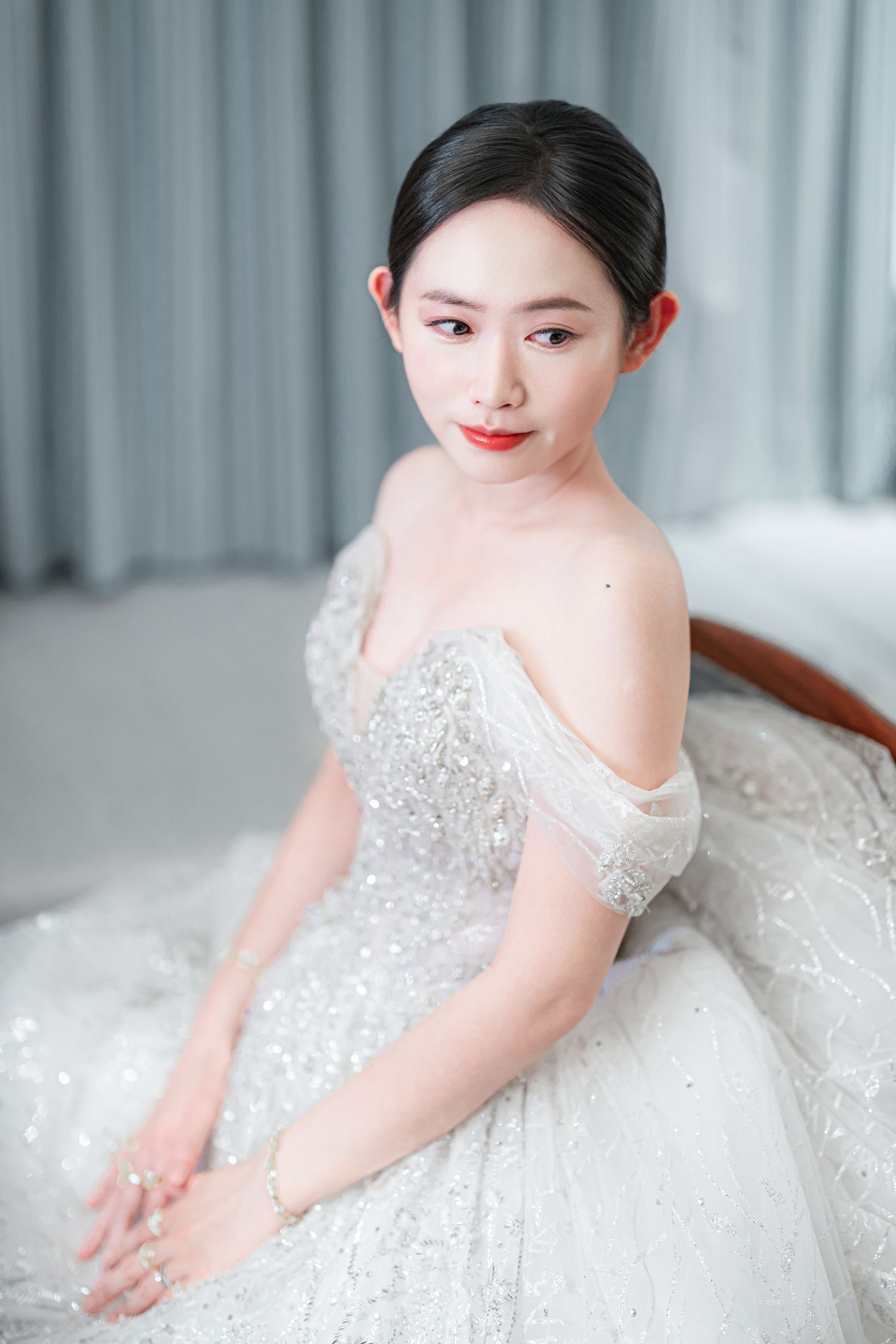 A bride in a sparkly white wedding dress with crystal embellishments sits gracefully in a light-filled room.