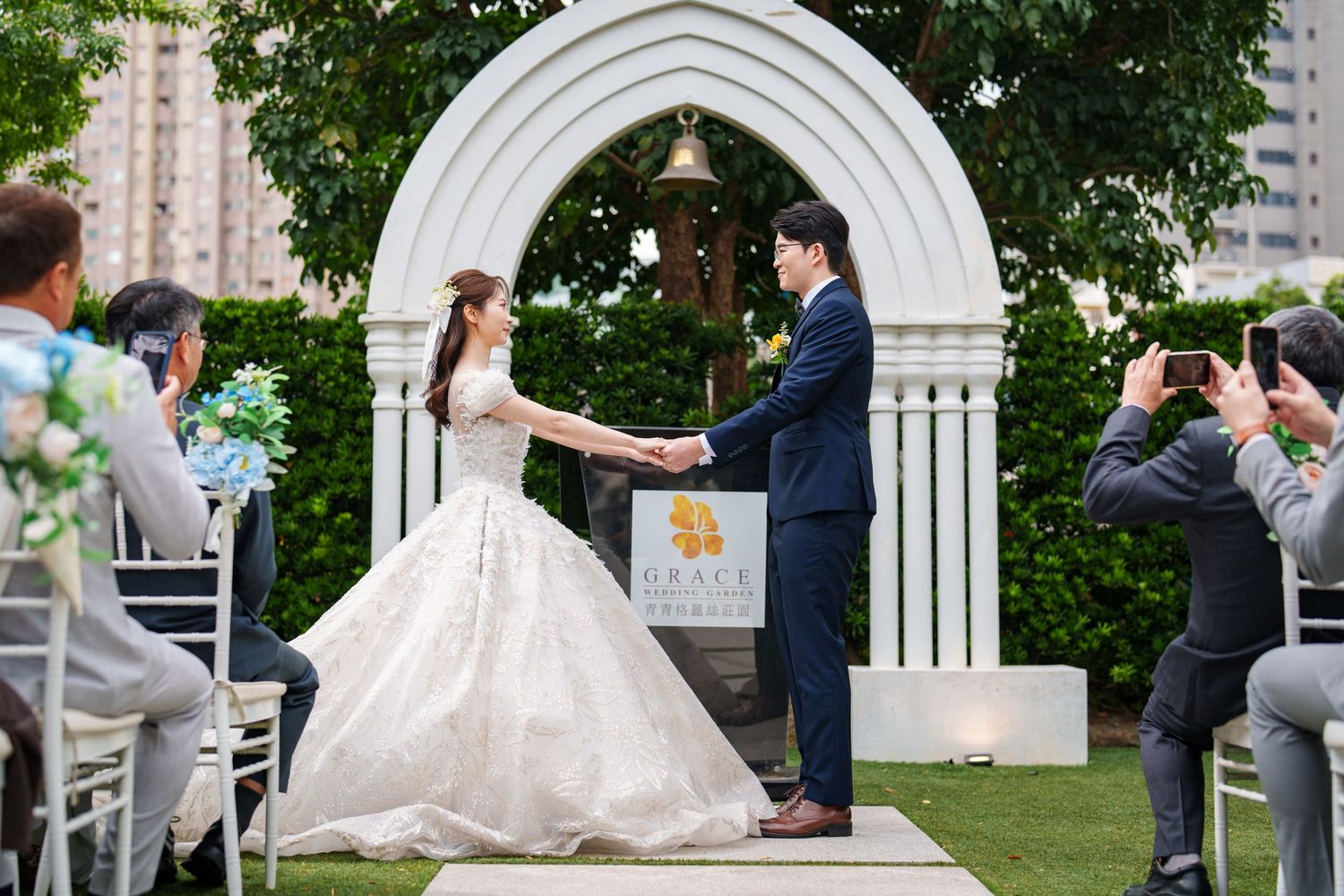 A romantic outdoor wedding ceremony takes place under a white decorative arch surrounded by lush greenery.