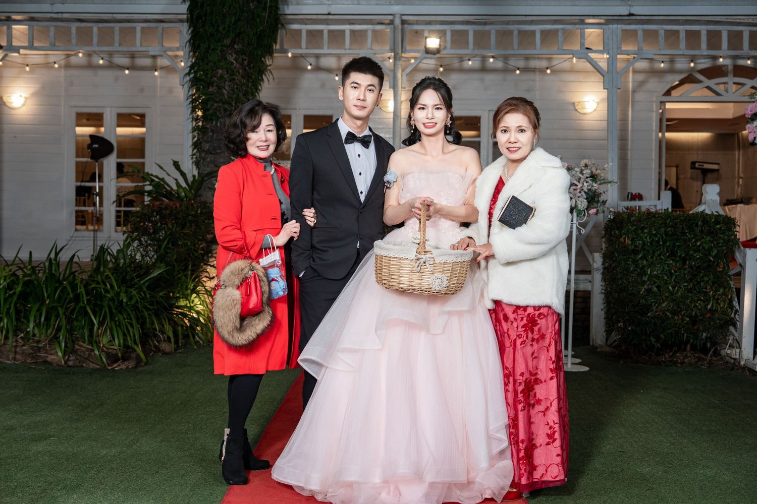 A formal wedding celebration outdoors with people in evening attire including a pink ballgown and red outfits at a venue.