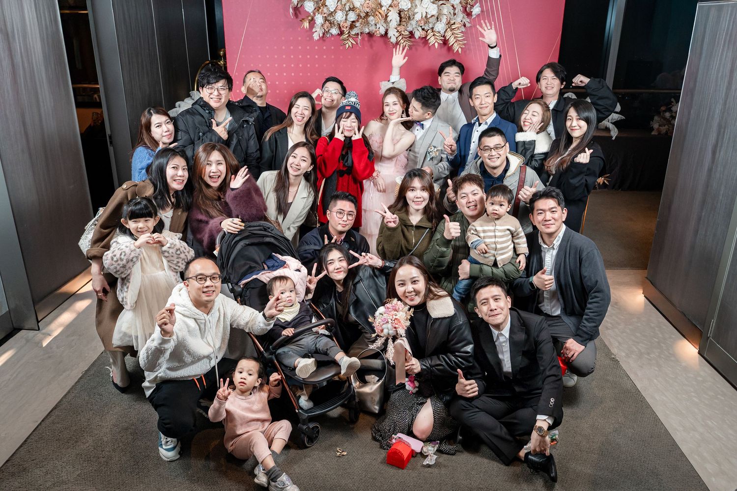 Large group of people gathered together indoors posing for a casual group photo against a pink wall with chandelier.