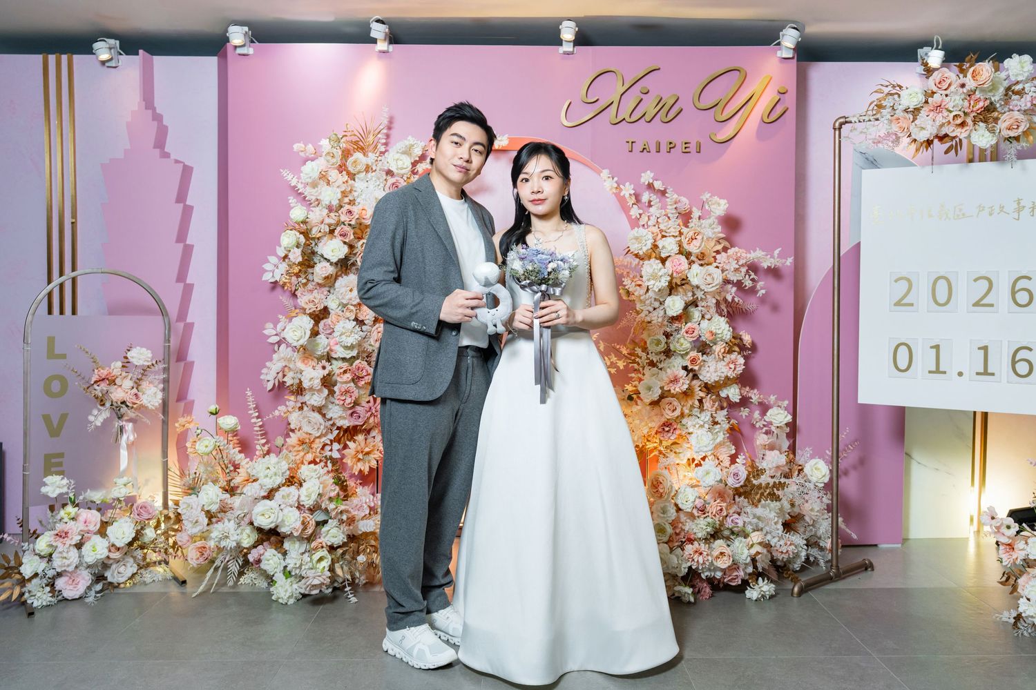 Wedding couple poses in front of floral arch with pink backdrop and elegant decor at romantic ceremony venue.