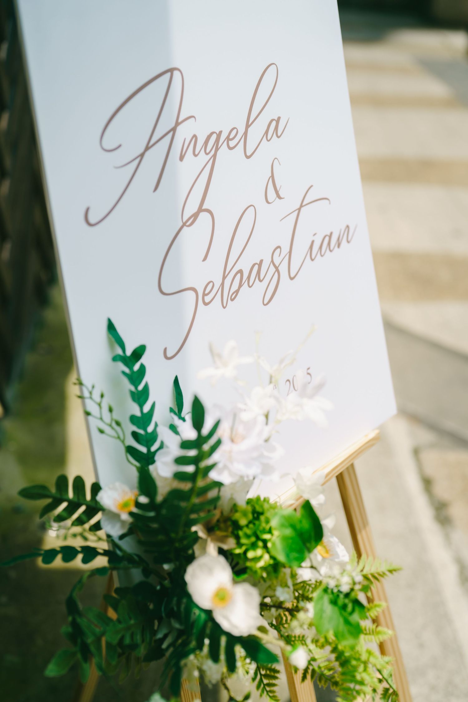 An elegant wedding sign with calligraphy names and decorative green foliage with white floral accents.